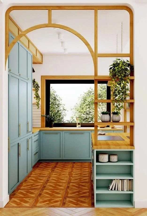 A kitchen with grey cabinets, a large window showing greenery outside, and wooden shelving with plants and dishes, separated by a decorative wooden partition.