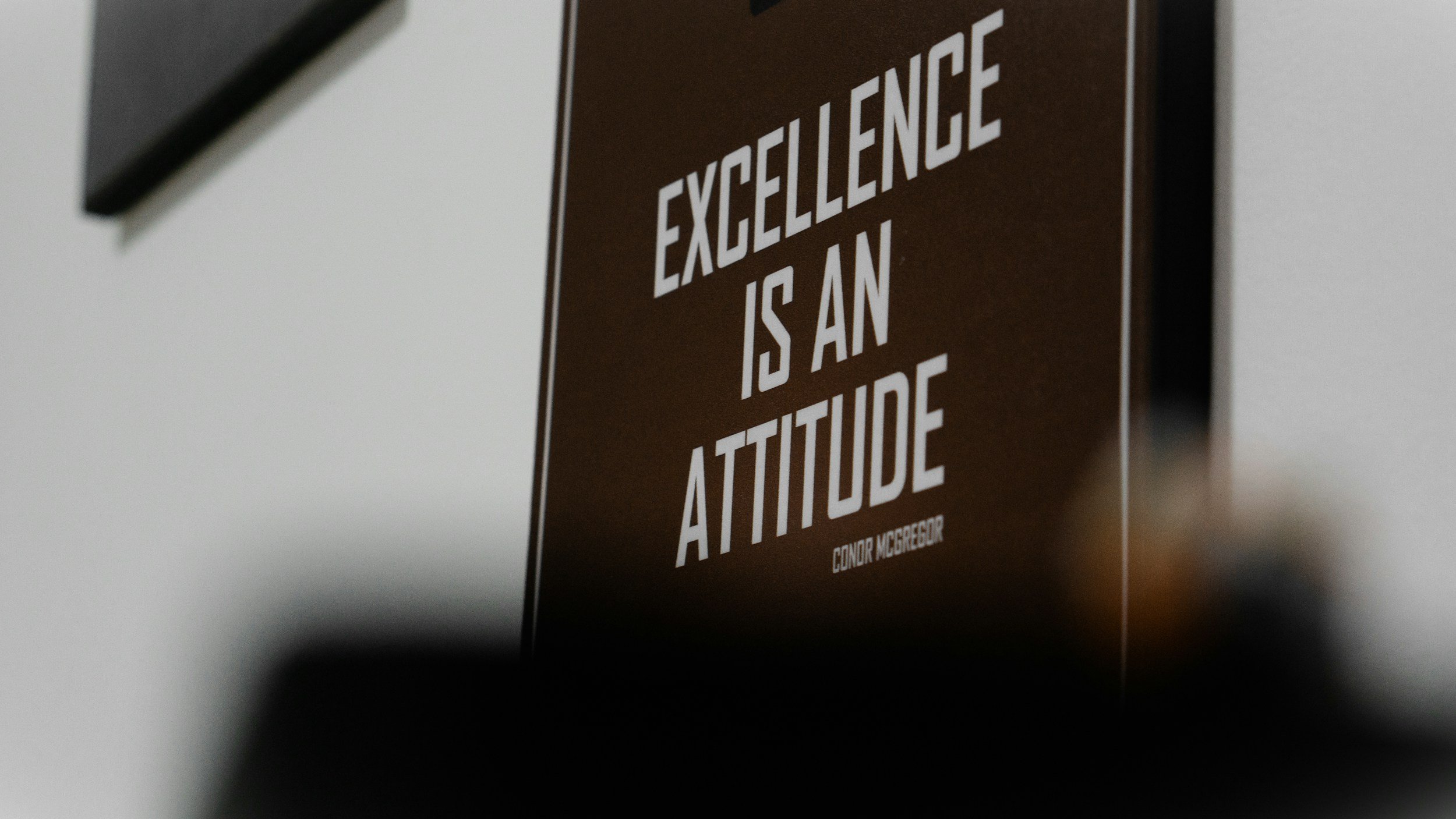 Close-up of a brown sign with white text that says 'Excellence is an Attitude' and a smaller text at the bottom with the name Conn McGregor.