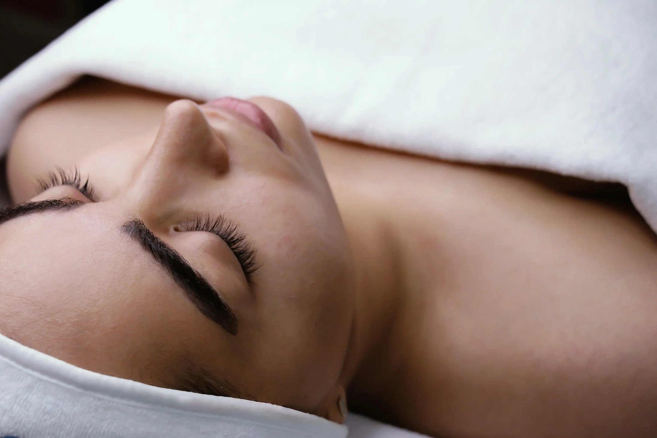 Close-up of a woman lying down with eyes closed, covered with a white towel or cloth.