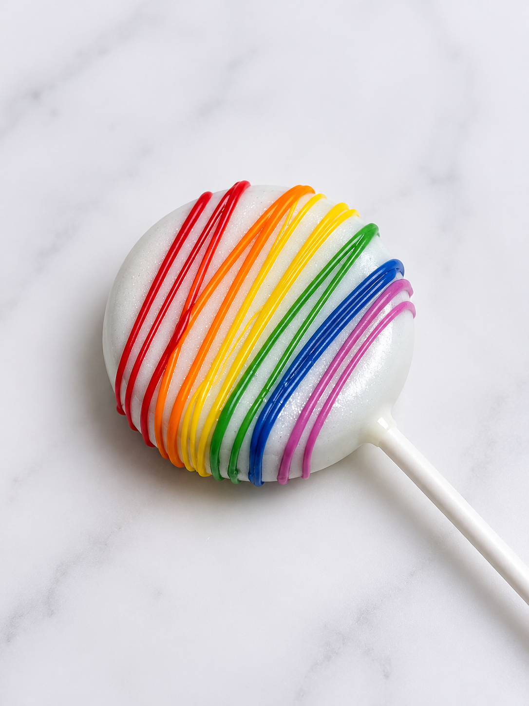 A rainbow-colored lollipop with white base and rainbow-colored candy coating, on a white marble surface.