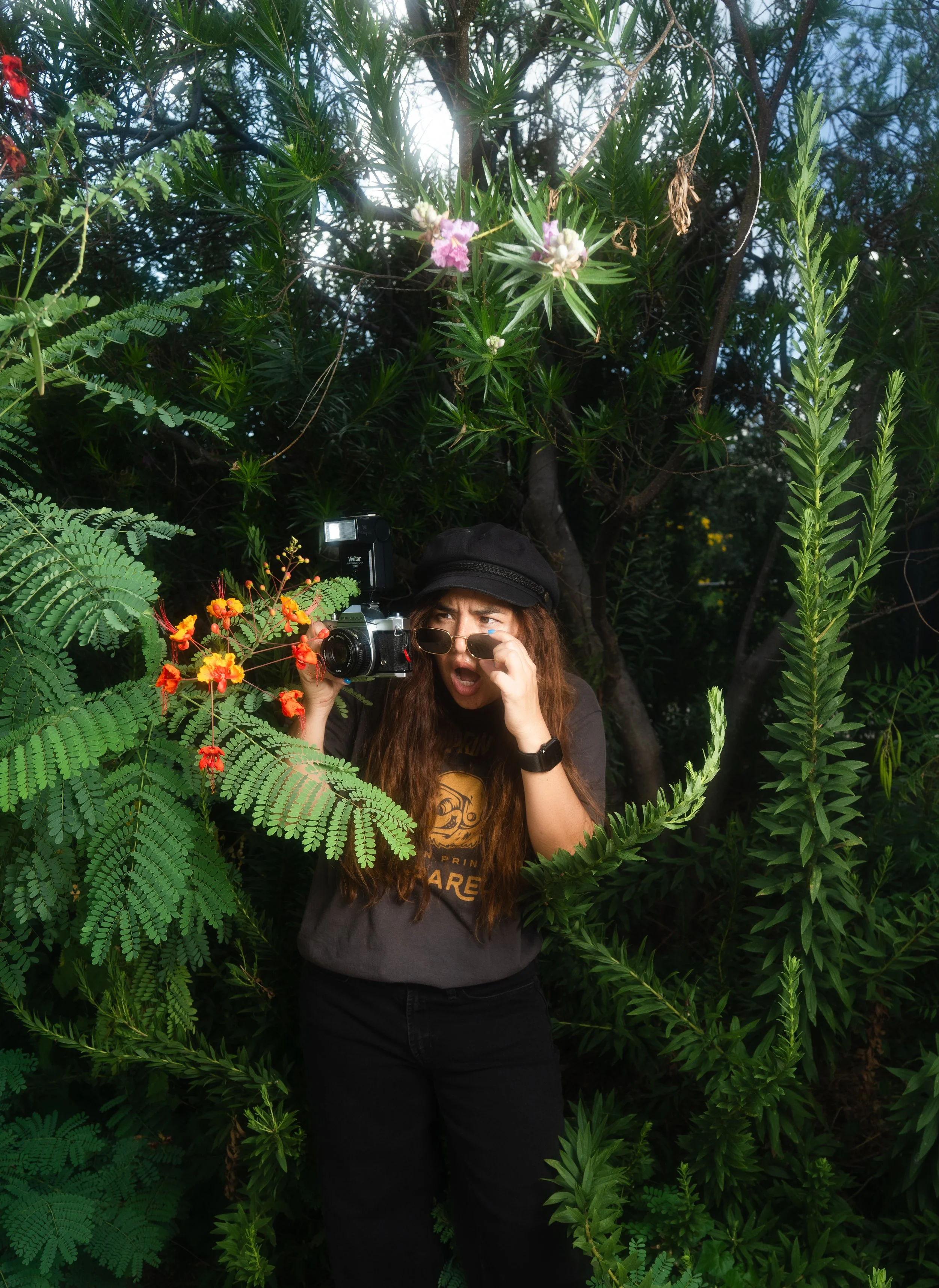 A woman with long brown hair and glasses taking a photo with a camera amidst lush green plants and trees, wearing a black hat, black pants, and a black shirt with a yellow graphic.