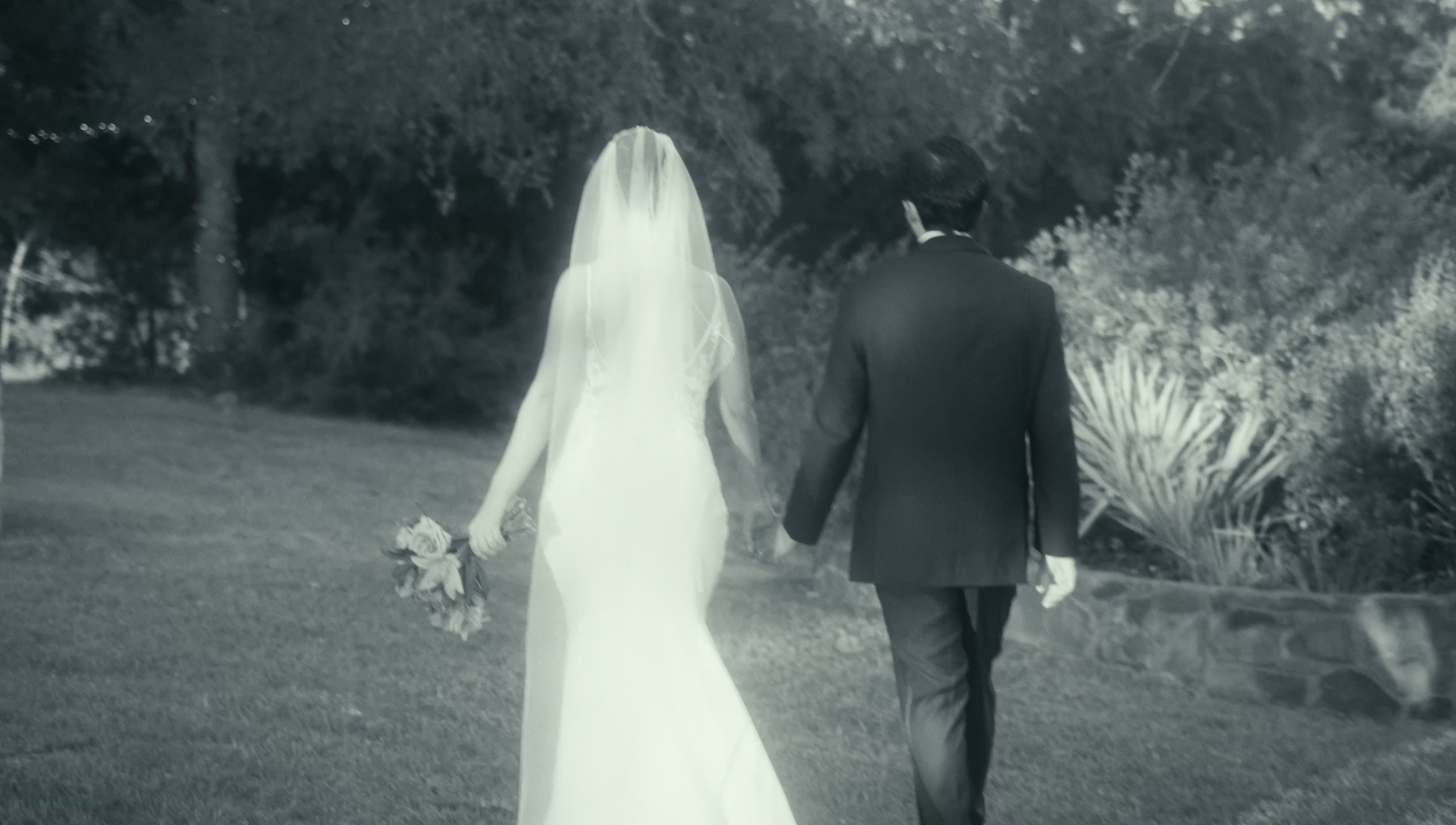 A black and white photo of a bride and groom walking away outdoors at night, with the bride in a white wedding dress and veil holding a bouquet of flowers, and the groom in a dark suit.