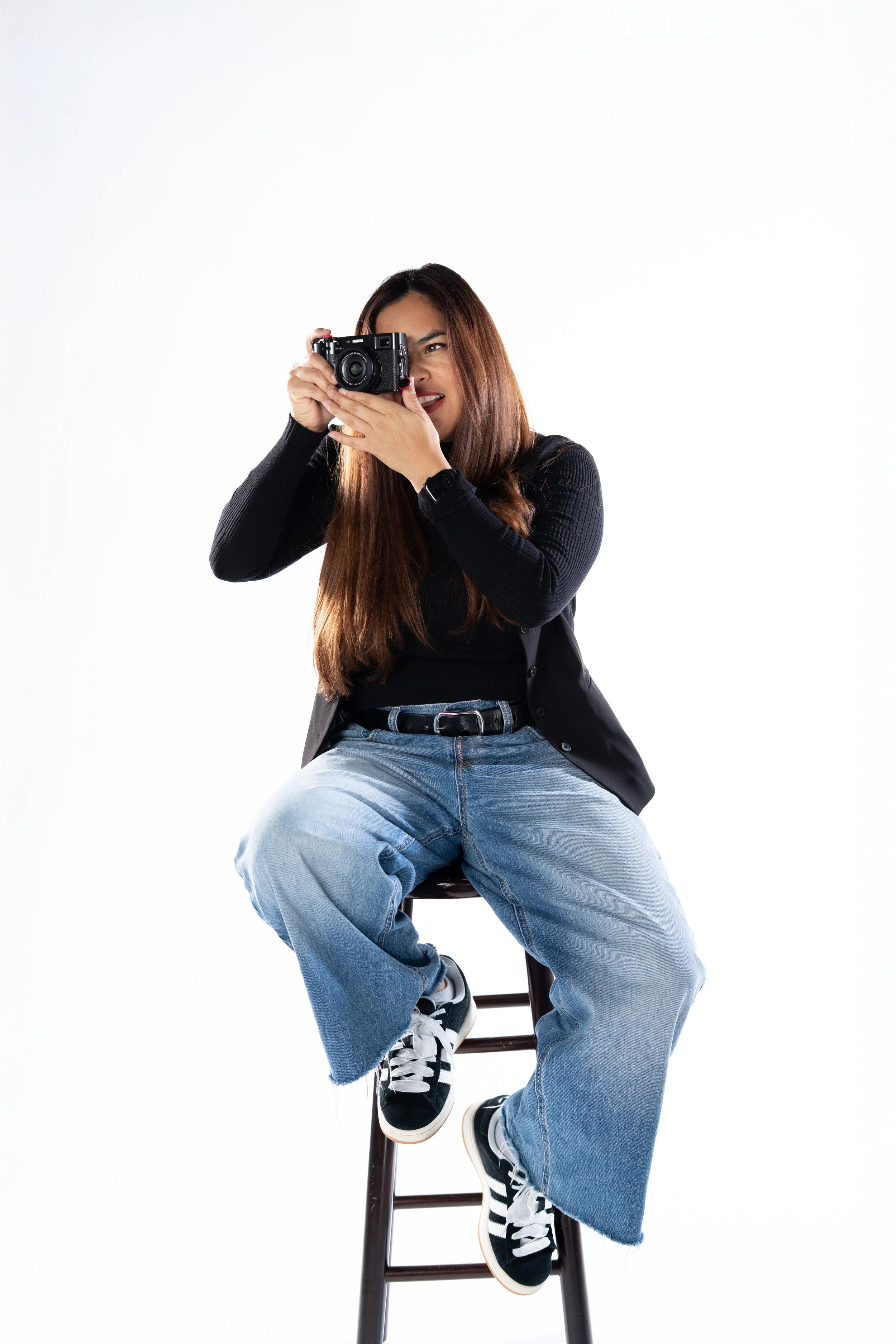 A young woman with long brown hair sitting on a tall wooden stool, holding a camera up to her eye, wearing a black jacket, blue jeans, and black and white sneakers. She is looking through the camera lens, with a white background.