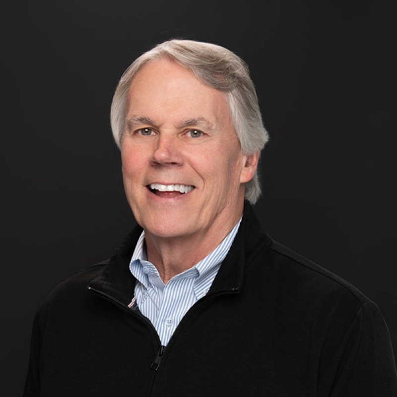 Portrait of a smiling middle-aged man with gray hair, wearing a black jacket over a blue and white striped shirt, against a dark background.