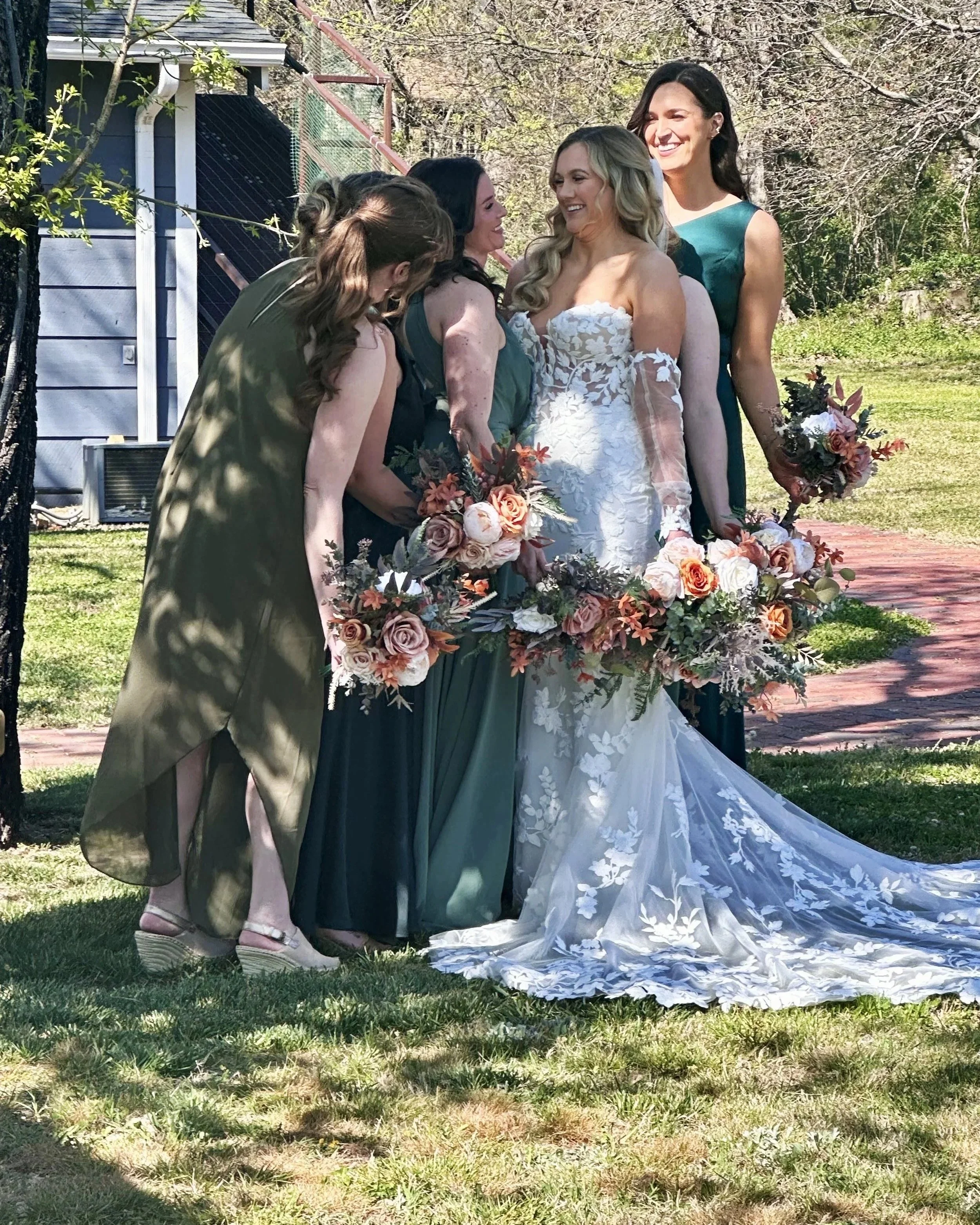 A bridal party of five women standing outdoors in a backyard, smiling and laughing, with greenery and trees in the background. The woman in the center is wearing a white wedding dress, while the others are wearing dark green dresses. They are holding