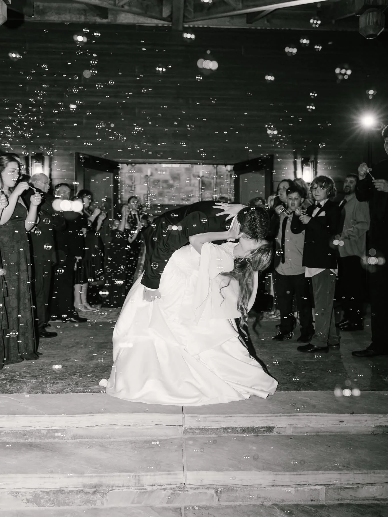 A wedding reception with a couple dancing closely, surrounded by guests taking photos and blowing bubbles, in a decorated indoor venue.