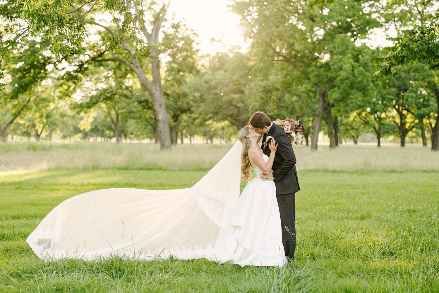 A bride and groom share a kiss in a grassy park with green trees, sunlight filtering through the leaves. The bride wears a white wedding dress with a long train, and the groom is in a black suit. She holds a bouquet of flowers.