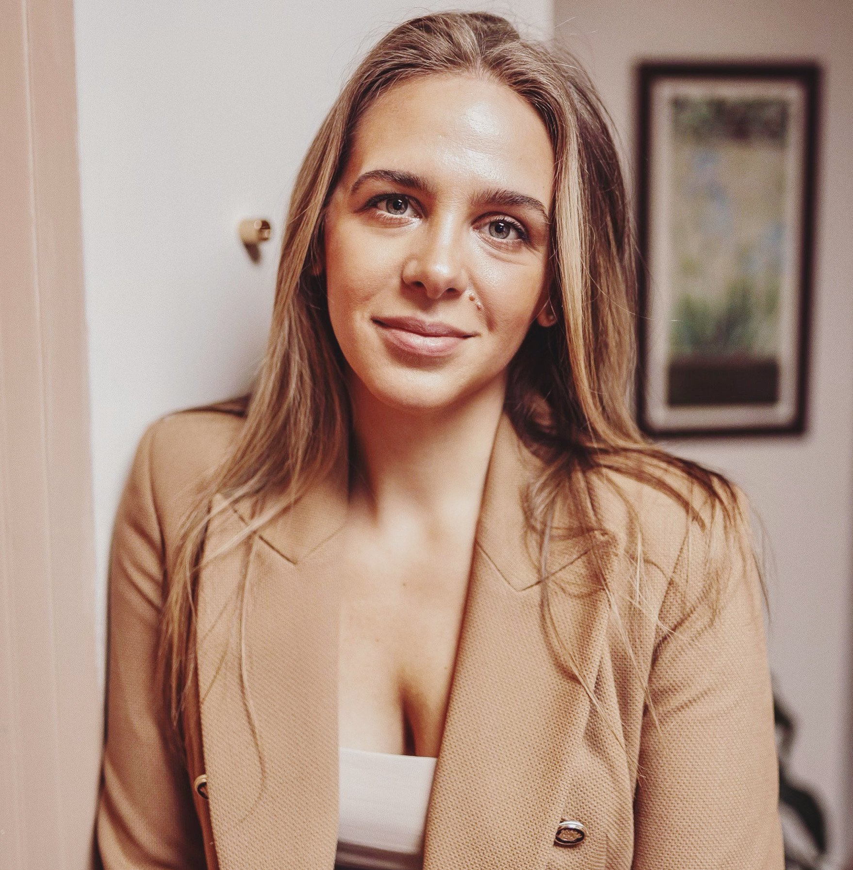 Close-up of a woman with light brown hair and blue eyes, smiling gently, wearing a beige textured blazer over a white top, with a framed picture in the background.