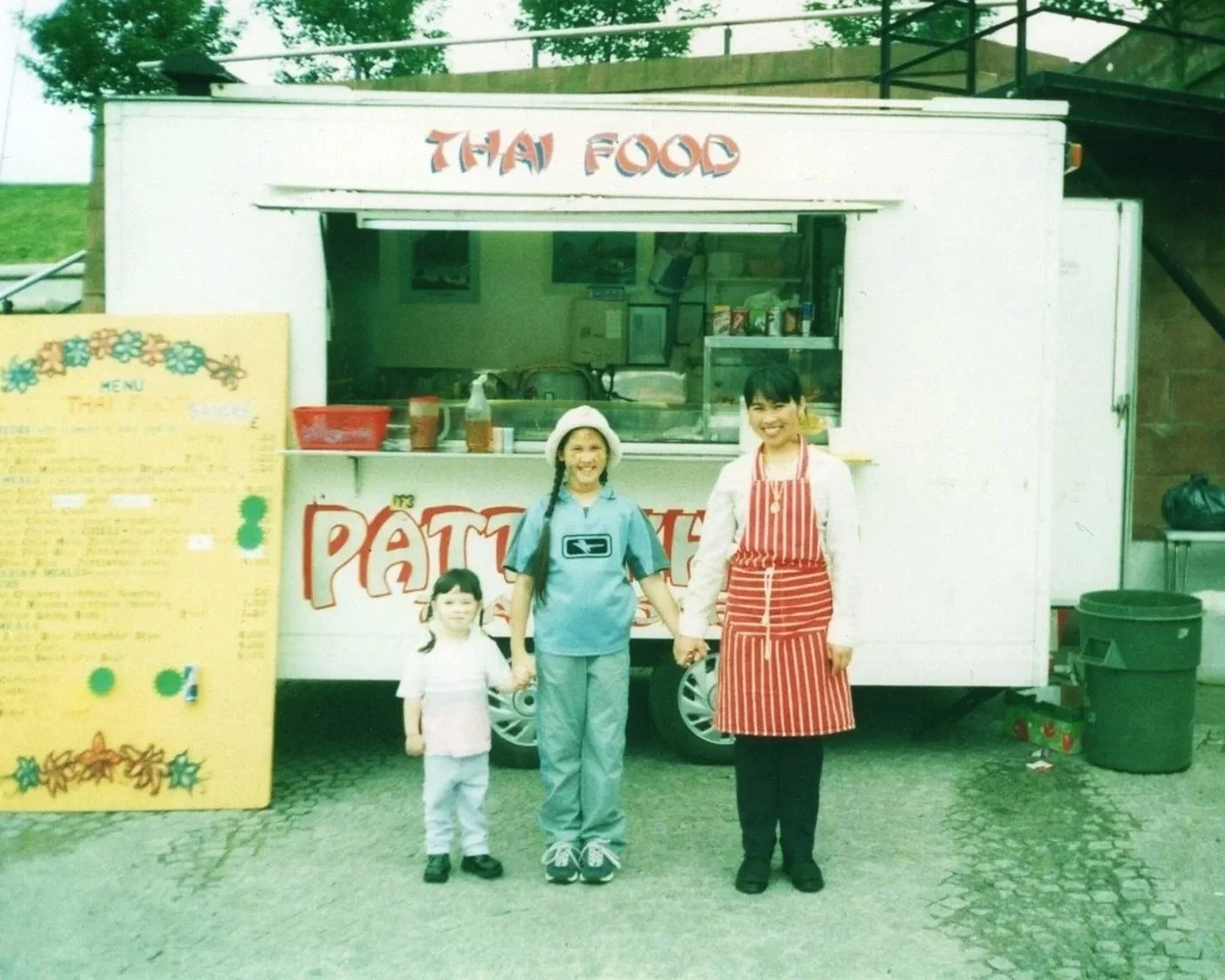 A family of three holding hands in front of a food stand labeled 'Thai Food.' The woman is wearing a red apron, and the children are wearing casual clothes. The stand has a menu board on the left side.