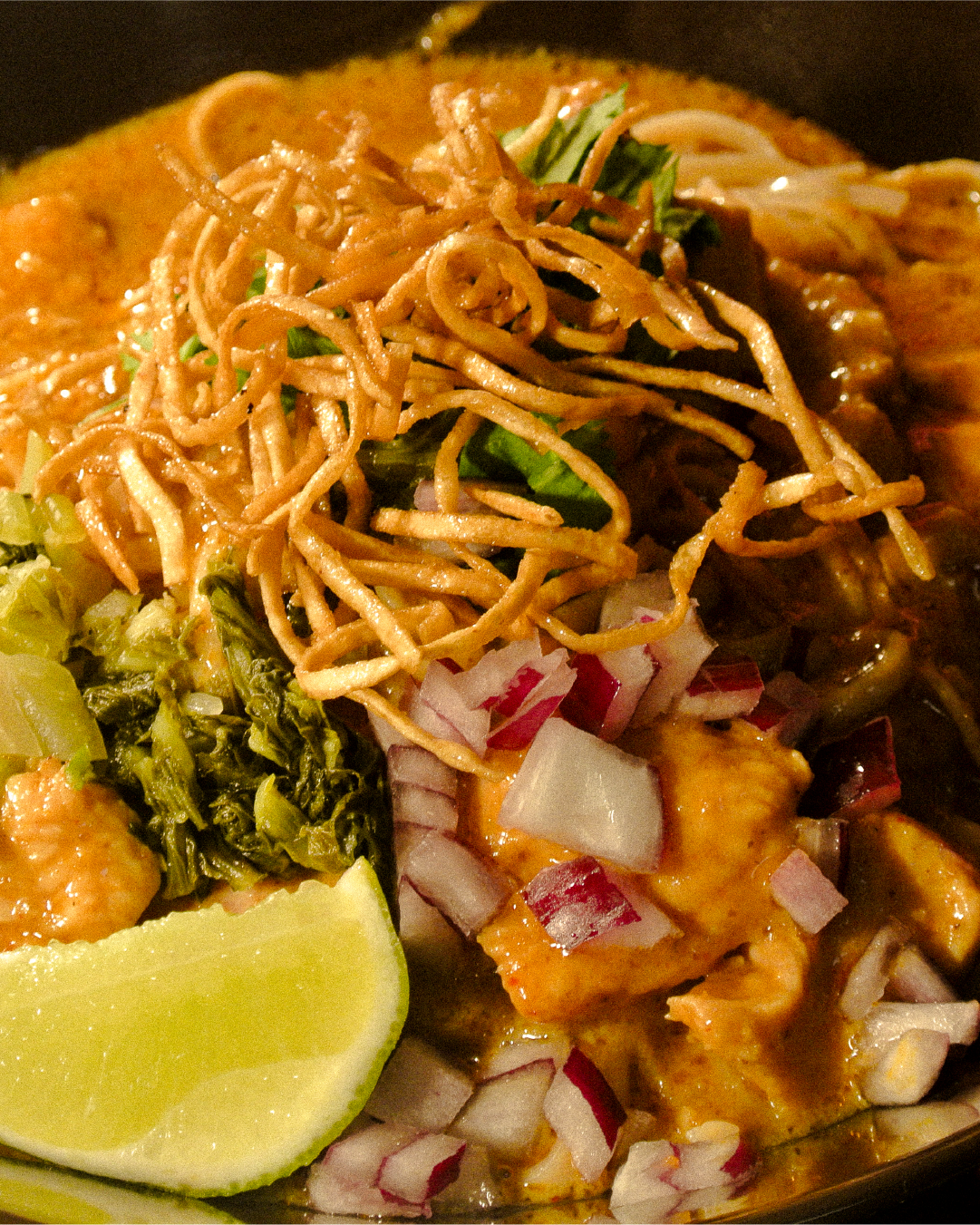 Close-up of a plate of Asian cuisine with noodles, chopped onions, chopped red cabbage, chopped greens, a lemon wedge, and curry.