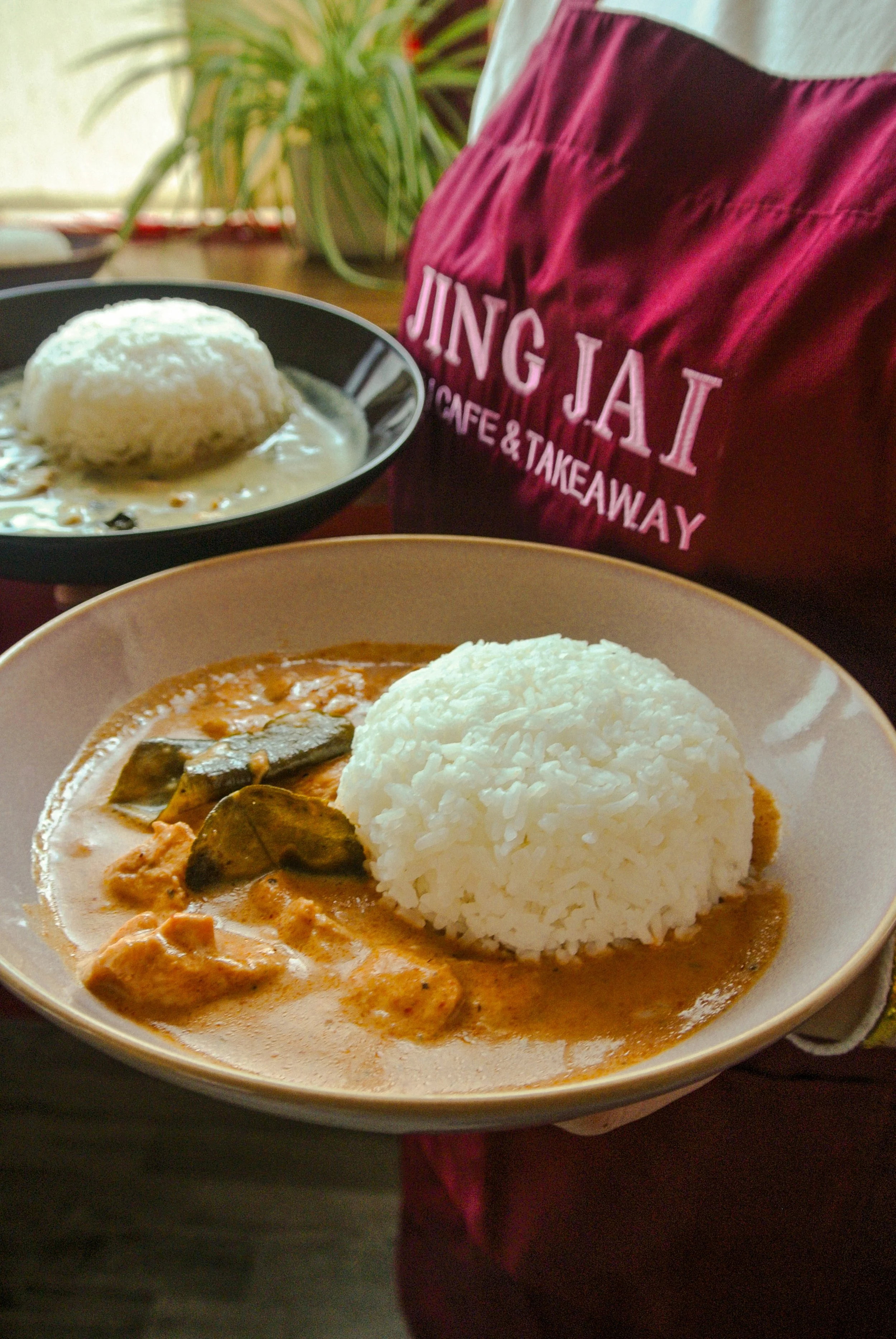 A plate of massaman curry with white rice next to it, accompanied by a bowl of thai green curry with white rice, in a restaurant setting with a person wearing a red apron that has the text 'JING JAI' and 'CAFE & TAKEAWAY' on it.