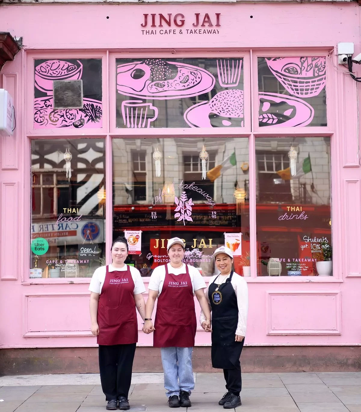 Three women standing in front of a pink Thai cafe and takeaway called Jing JAI, holding hands and smiling. The cafe has a large window with illustrations of Thai food and drinks, and the women are wearing aprons and caps, two in red and one in black.