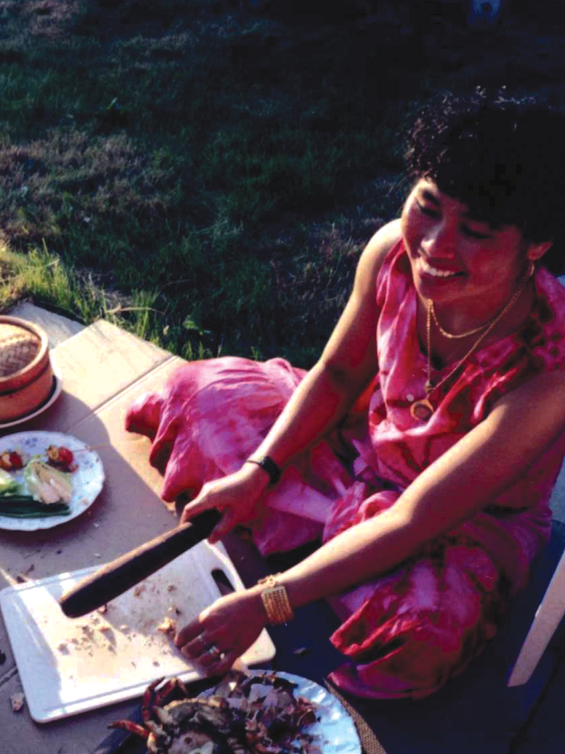 A woman sitting outdoors on the grass is slicing food on a cutting board. She is wearing a pink dress, gold jewelry, and has short curly hair. There are plates with food and bowls on the table beside her.
