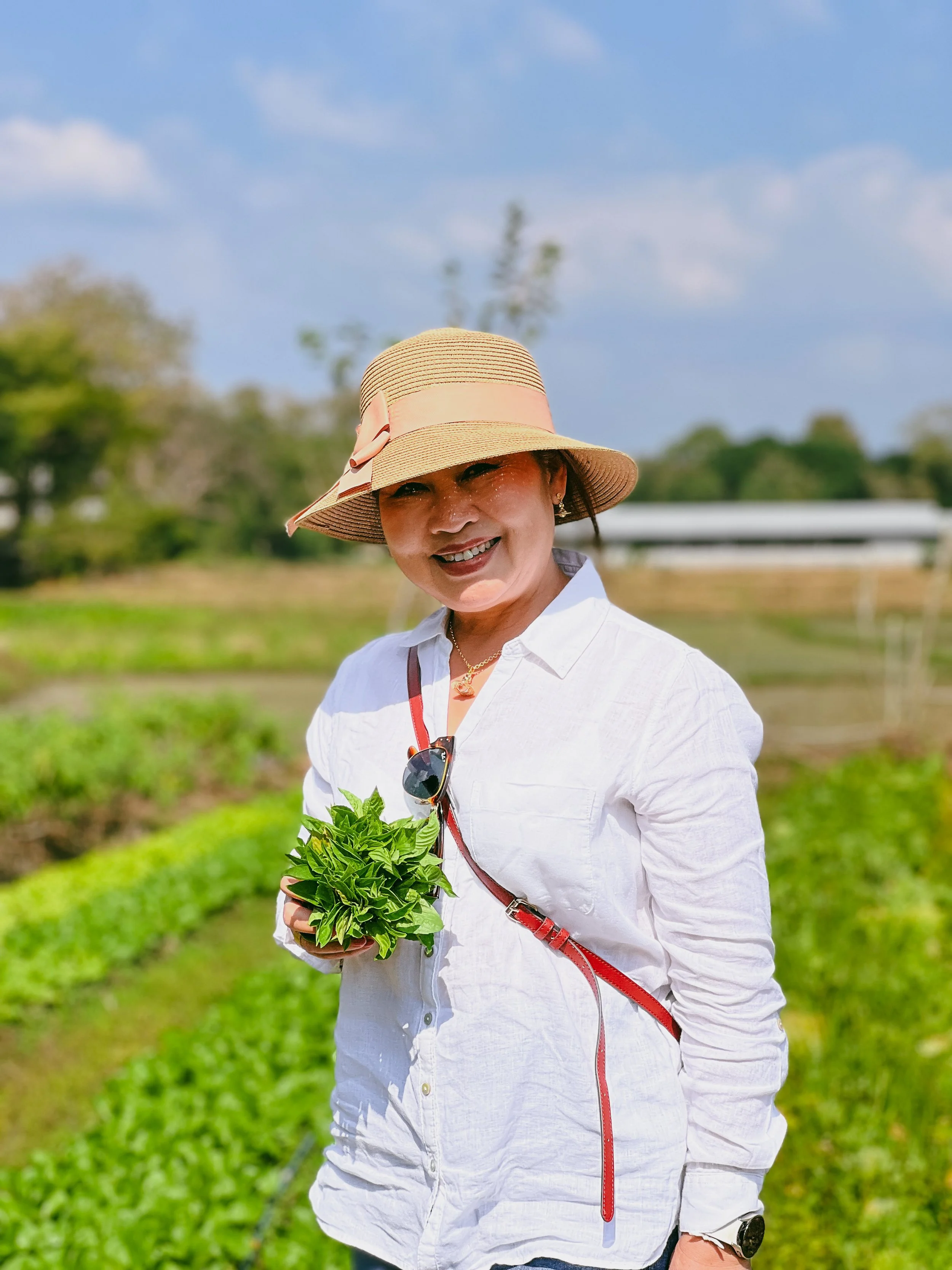 Woman wearing a straw hat with a pink ribbon, white shirt, and sunglasses hanging on her shirt, smiling and holding a bunch of fresh green herbs in an outdoor garden with green plants and a blue sky in the background.