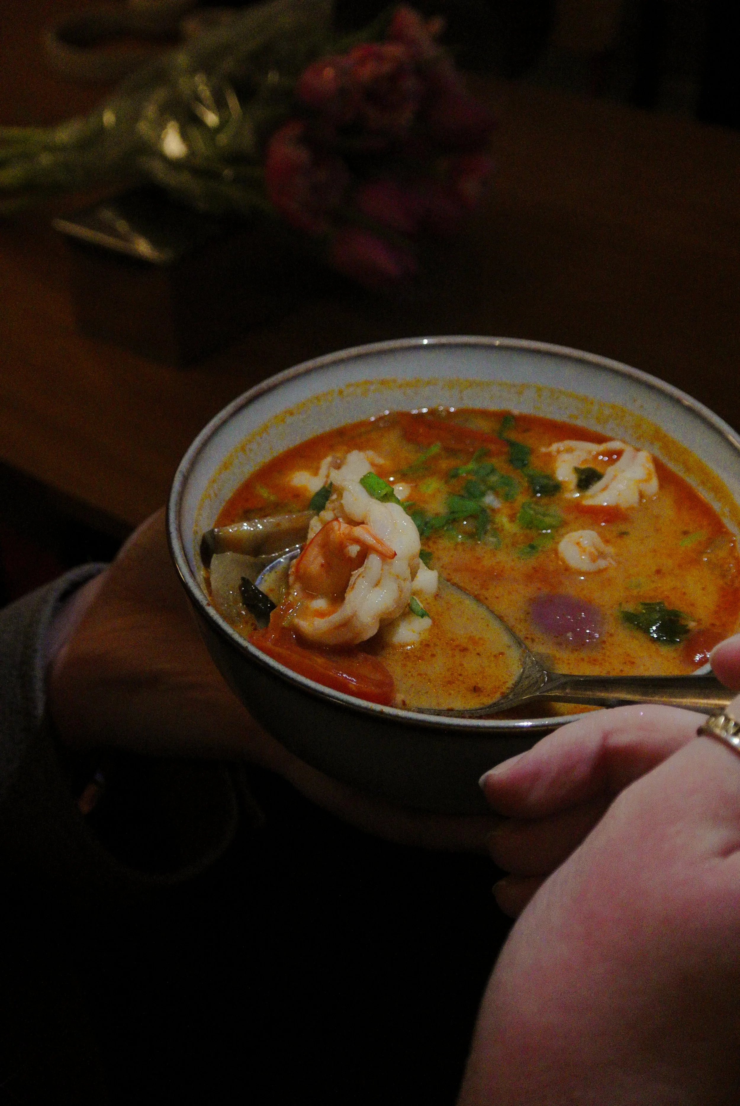 Close-up of a bowl of spicy seafood soup with shrimp, vegetables, and herbs, held by a person's hand, with a blurred background of a table and flowers.