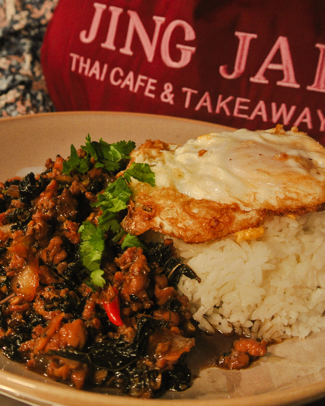 Close-up of a plate with rice, a fried egg, and stir-fried minced meat with vegetables, garnished with cilantro. In the background, a red bag with white text that reads 'JING JAI THAI CAFE & TAKEAWAY.'