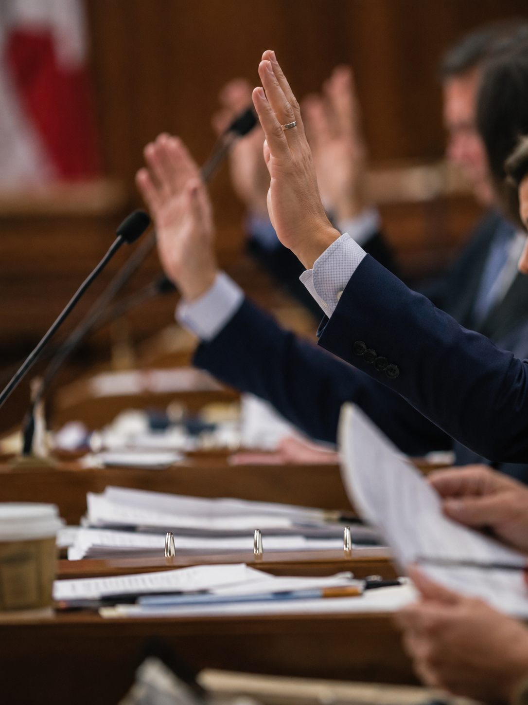 People in suits raising their hands during a meeting or hearing, with documents and a microphone on the table.