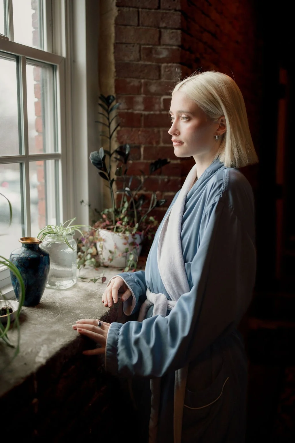 A blonde woman looking out a window on a cloudy day, standing by a windowsill with potted plants and vases, next to a brick wall.