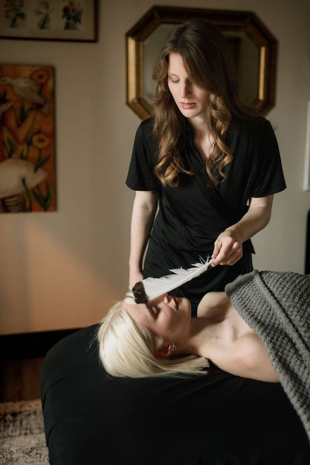 A woman with long, wavy brown hair dressed in a black outfit is holding a large white feather above the face of another woman with platinum blonde hair lying on a black surface, who is partially covered with a grey blanket.