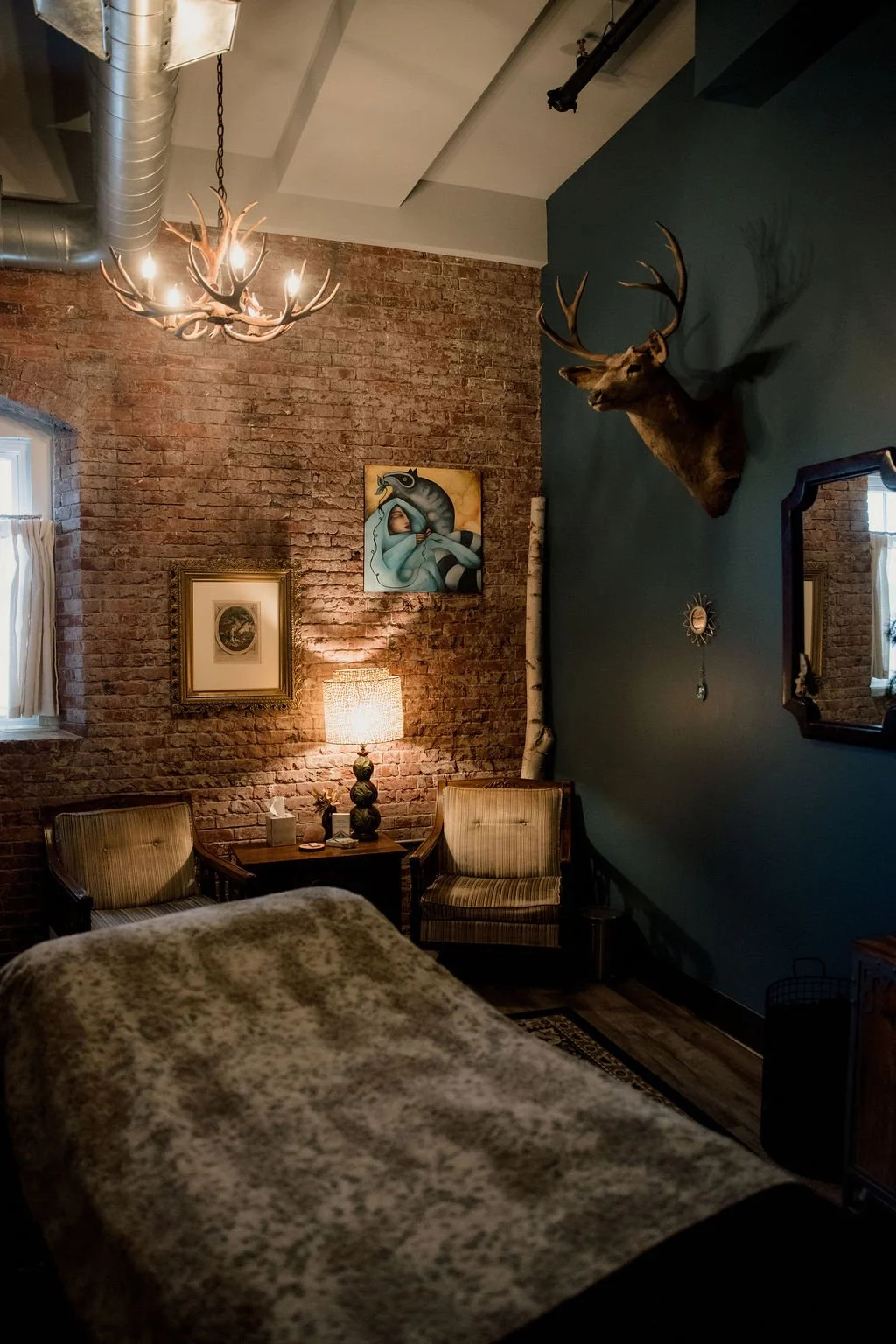 Cozy room with a brick wall, mounted deer head, antique armchairs, a small table with a lamp, artwork, and a mounted moose skull with antlers. Natural light coming through a window with white curtains.