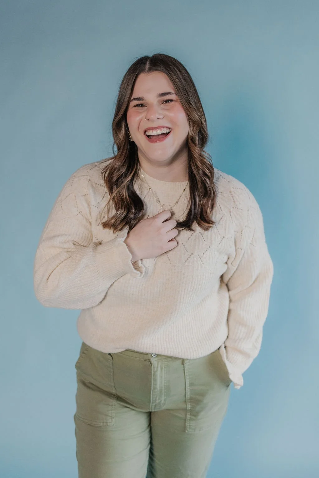 A young woman with long wavy brown hair, smiling and laughing, standing against a light blue background wearing a cream-colored knit sweater and light green pants.