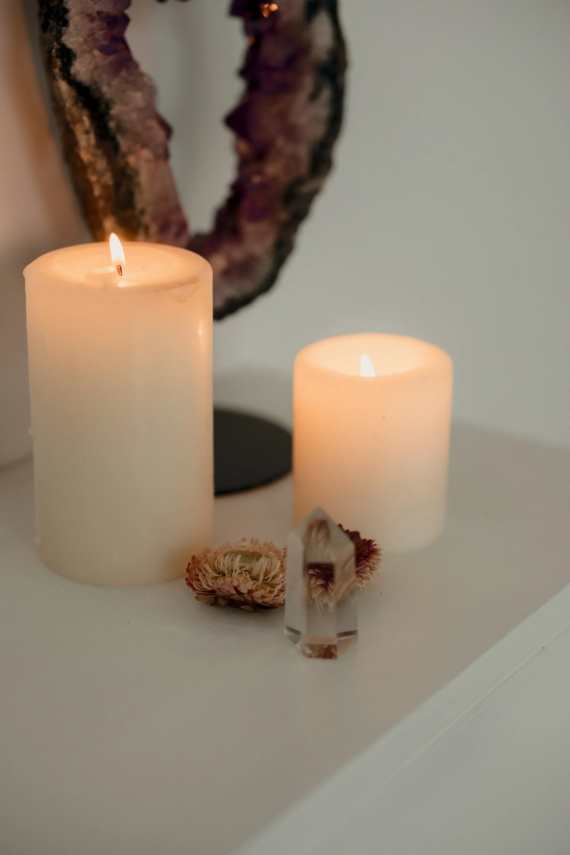 Two white candles burning on a surface, with decorative dried flowers and a clear crystal in front, and a round, textured object in the background.