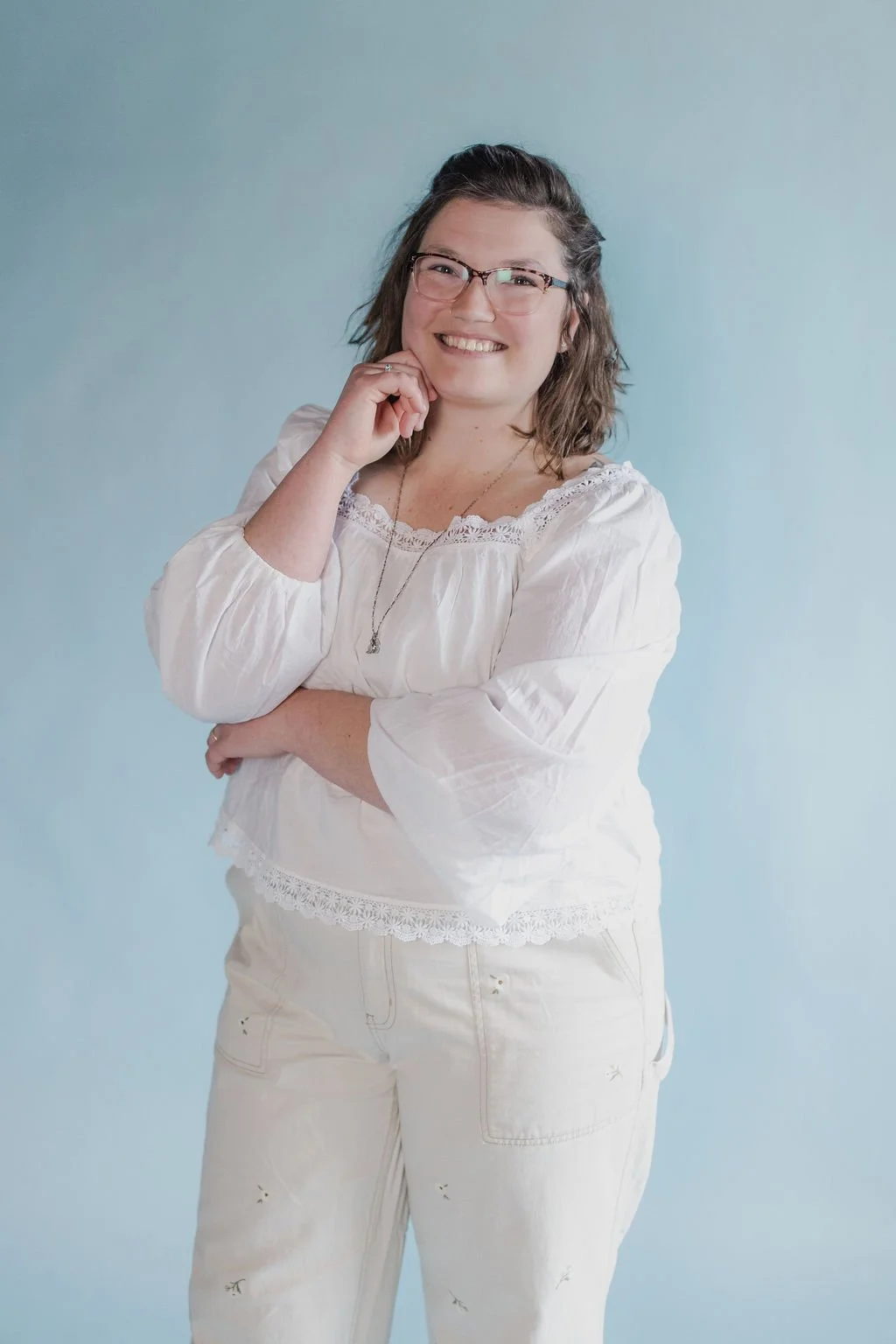 A woman with glasses, smiling, wearing a white blouse and white pants, standing against a light blue background.