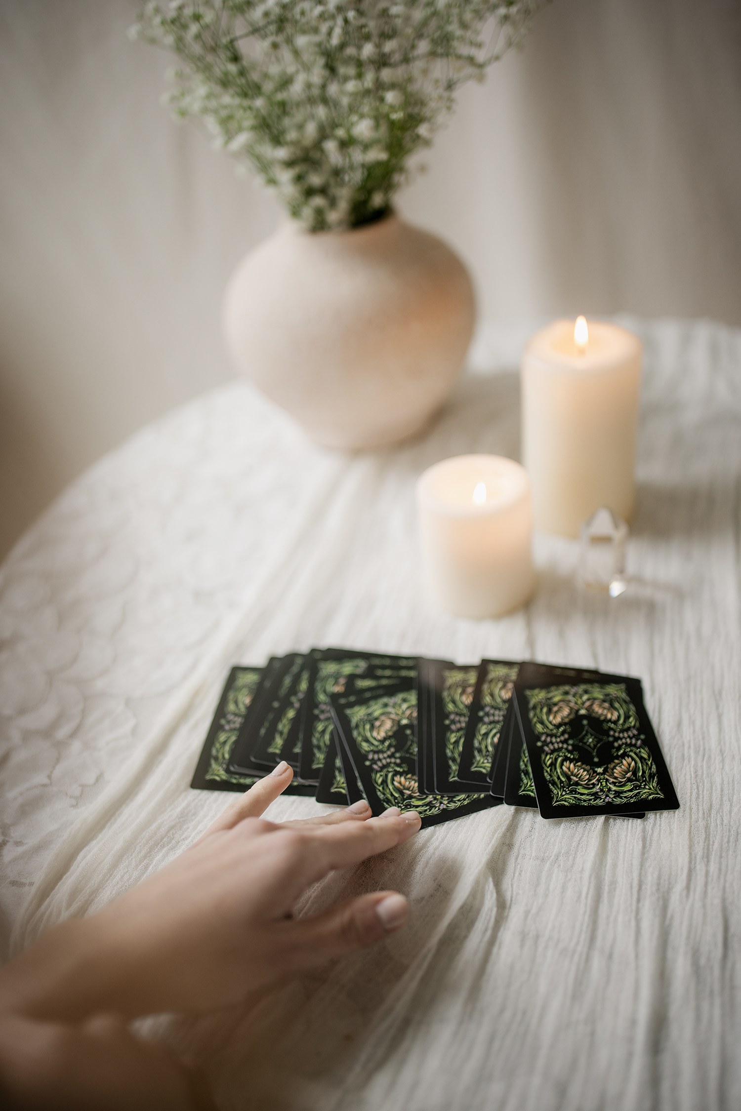 Person using tarot cards on a white table, with a large white candle, a smaller candle, a house-shaped figure, a vase with white flowers, and tarot card images.