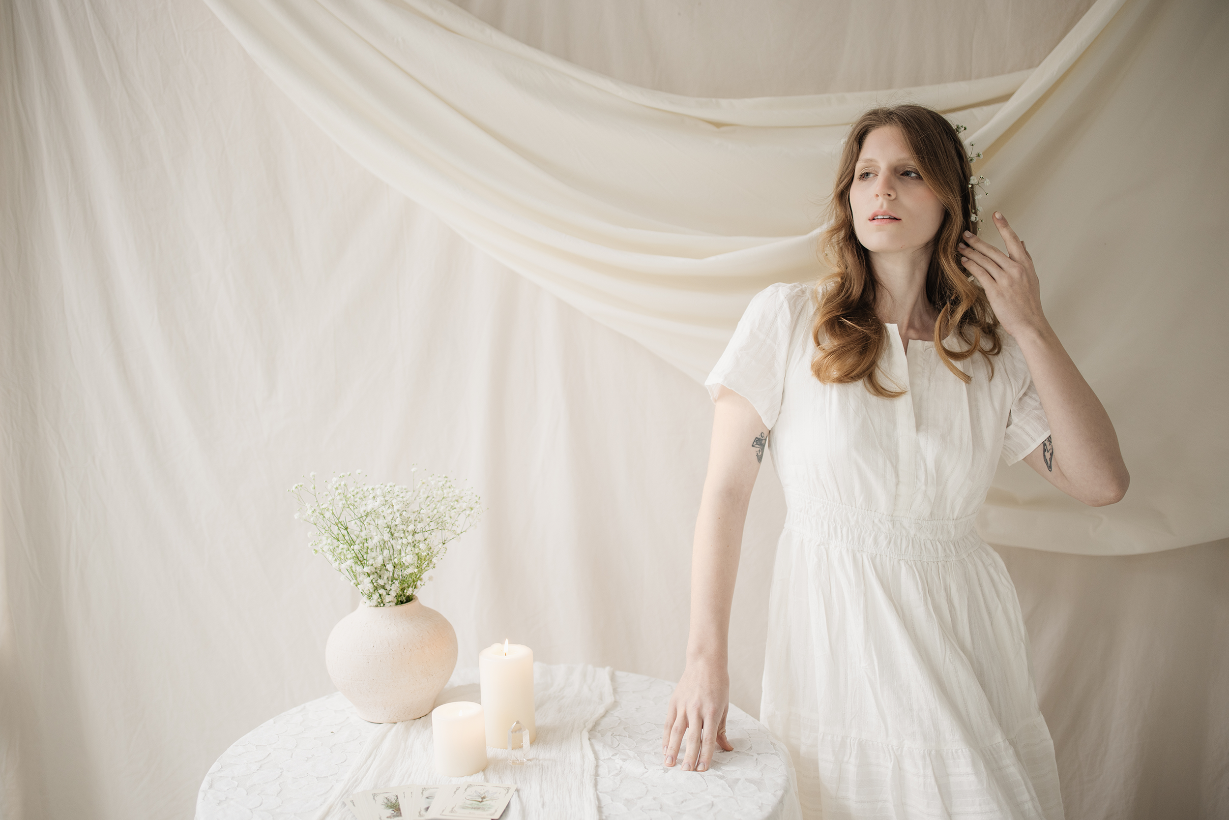 A woman with tattoos wearing a white dress, standing indoors with a cream-colored draped fabric background, near a table with candles, a vase with white flowers, and tarot cards.