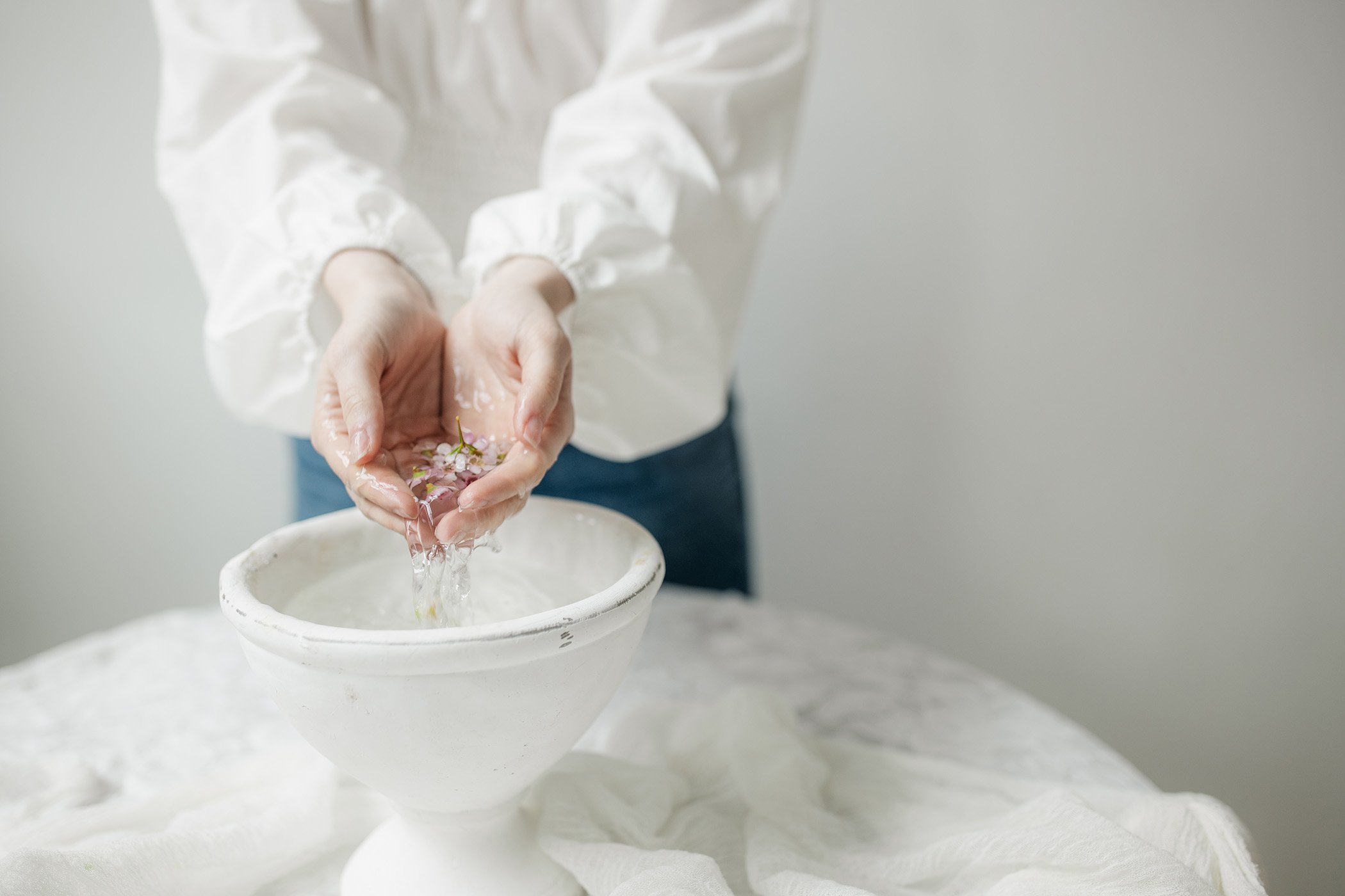 Close-up of a person in a white long-sleeve shirt pouring water from their hands, with pink flowers in their palms, into a bowl, on a white surface against a plain gray background.