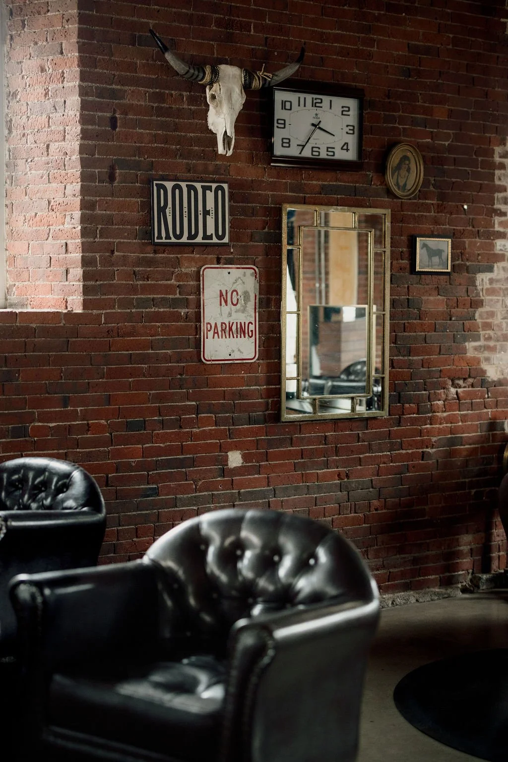 Interior of a rustic room with a brick wall decorated with a mounted animal skull with horns, a vintage clock, a mirror, and framed pictures. There are black leather chairs in the foreground.