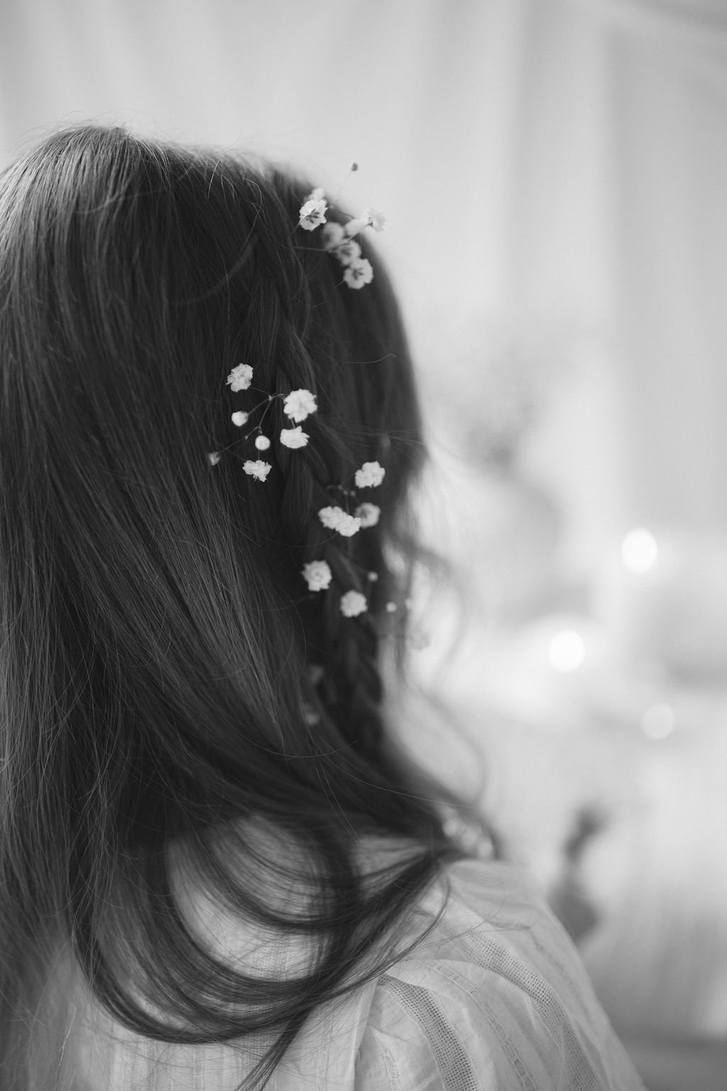 Black and white photo of a person with wavy hair decorated with small flowers, facing away from the camera.