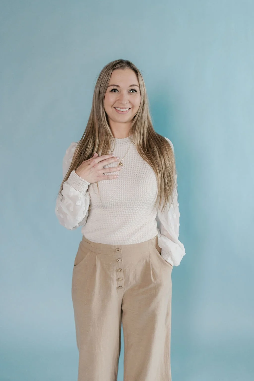 A young woman with long blonde hair smiling at the camera, wearing a cream-colored sweater with puffed sleeves and beige high-waisted pants, standing against a light blue background.