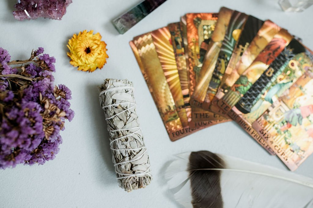 A bundle of smudging sage, a white feather, several tarot cards, a purple flower bouquet, and some dried yellow flowers on a light-colored surface.