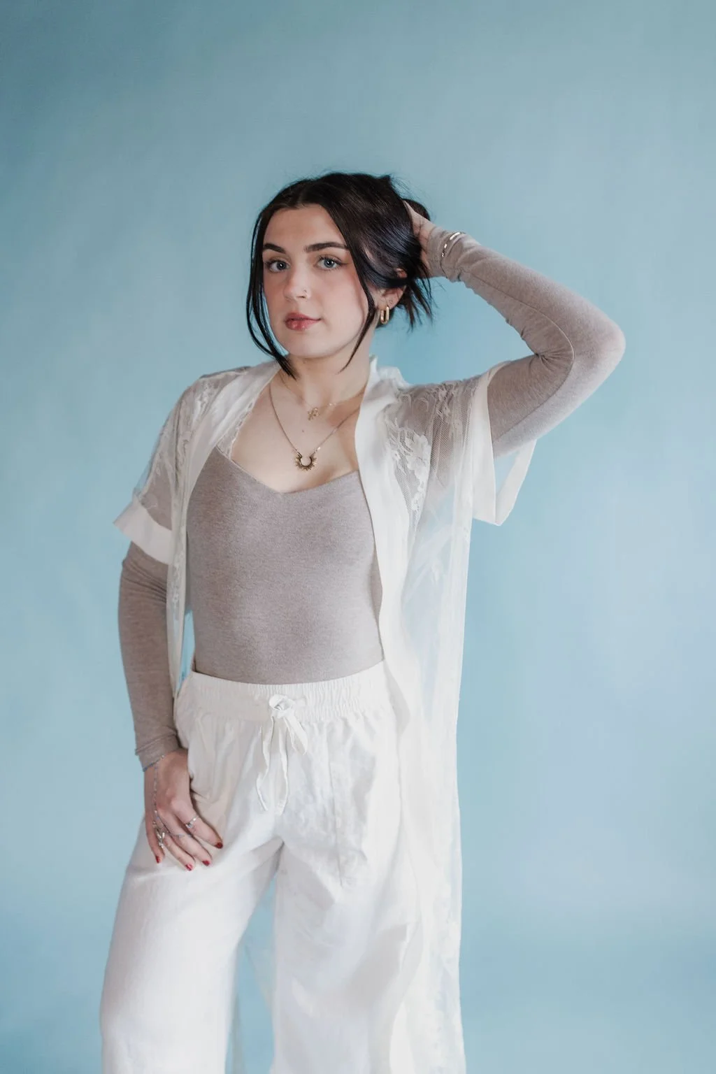 A young woman with shoulder-length dark hair, wearing a beige tank top, white pants, and a sheer white cover-up with lace sleeves, standing against a light blue background. She has jewelry including earrings, a necklace, and rings, and is posing with one hand behind her head.
