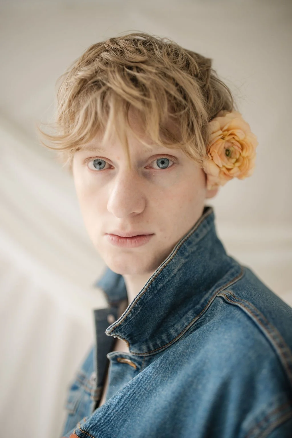 A young person with short, tousled red hair and blue eyes, wearing a blue denim jacket and a yellow-orange flower behind their ear, looking directly at the camera with a neutral expression.