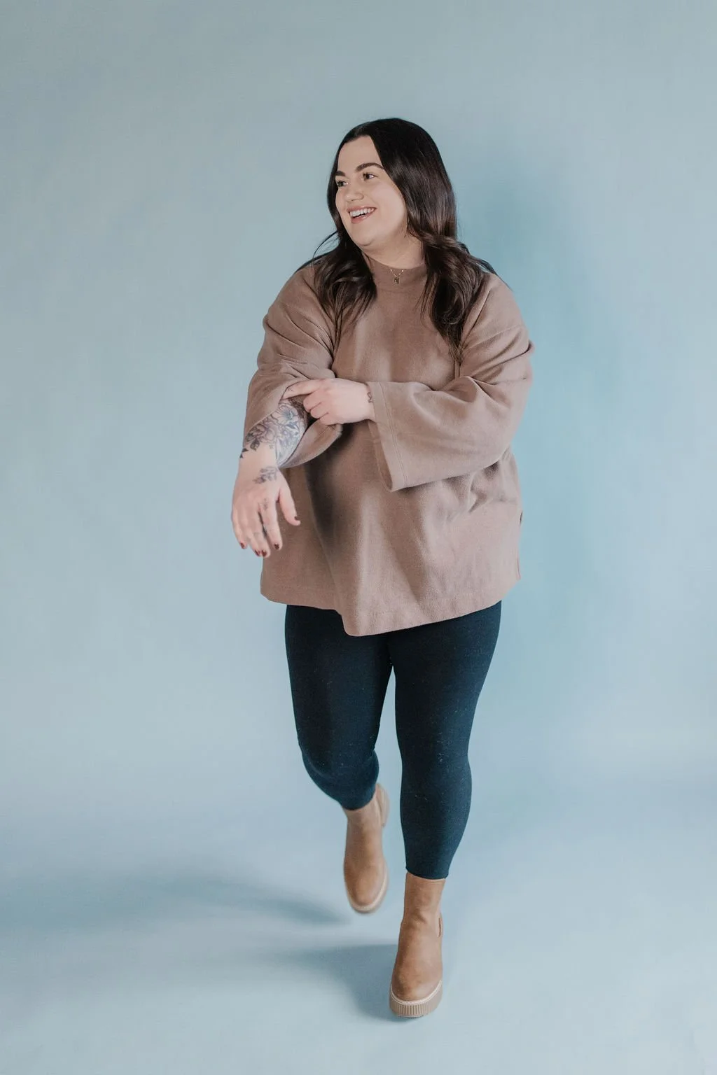 A woman with long dark hair, wearing a beige oversized sweatshirt, black leggings, and beige ankle boots, is smiling and running against a plain mint green background.