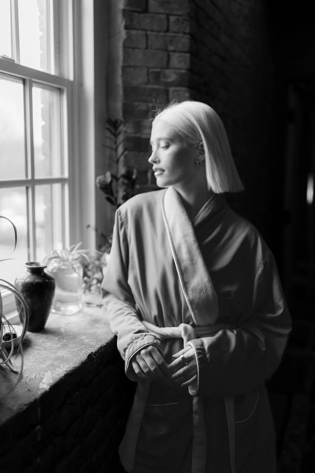 A woman with light hair, wearing a satin robe, looking out a window with plants and vases on the windowsill, in a brick-walled room, black and white photo.