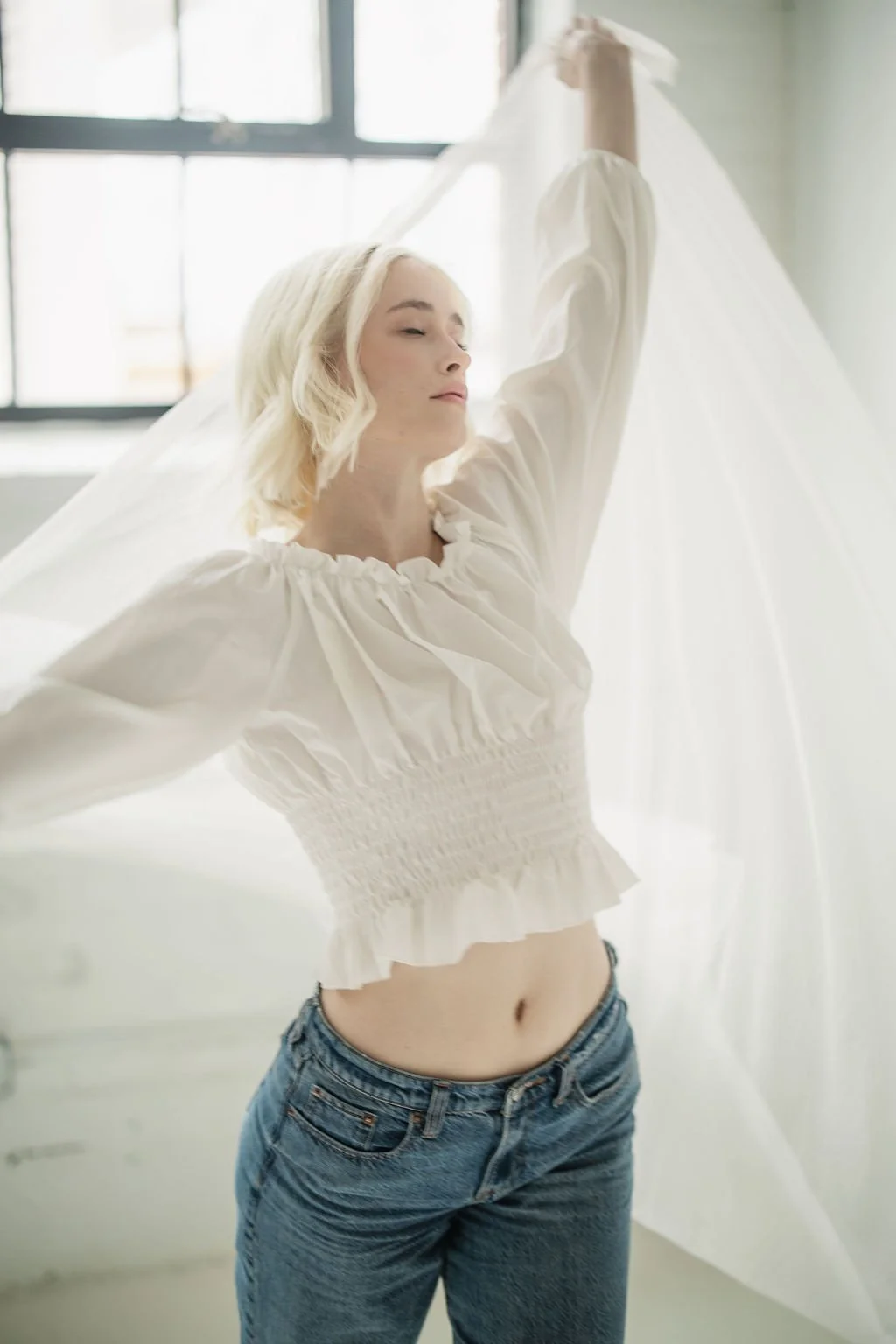 A woman with platinum blonde hair and fair skin is standing near a window, holding a sheer white fabric above her head, with her eyes closed and a serene expression. She is wearing a white, puff-sleeved, smocked crop top and blue jeans.