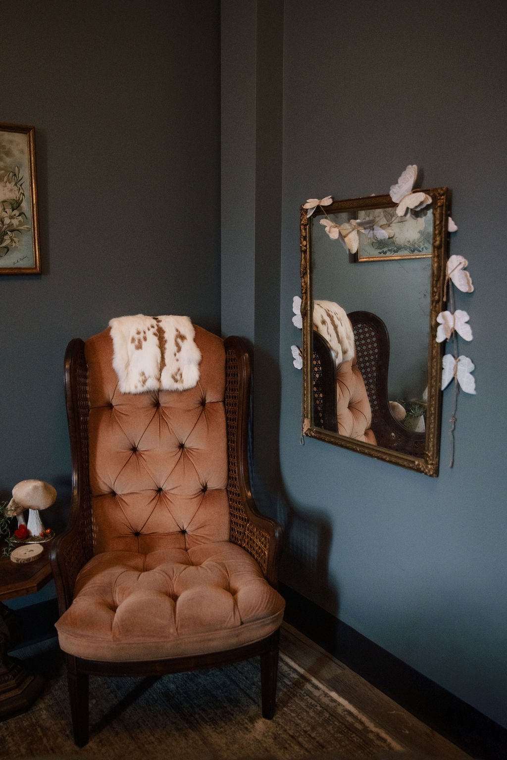 A vintage pink velvet armchair with tufted design and wooden frame, draped with a white fur throw, is placed in a room with dark teal walls. A mirror with a decorative frame hangs on the wall, adorned with butterfly-shaped ornaments. A small side table with a mushroom-shaped lamp and decorative items is nearby.