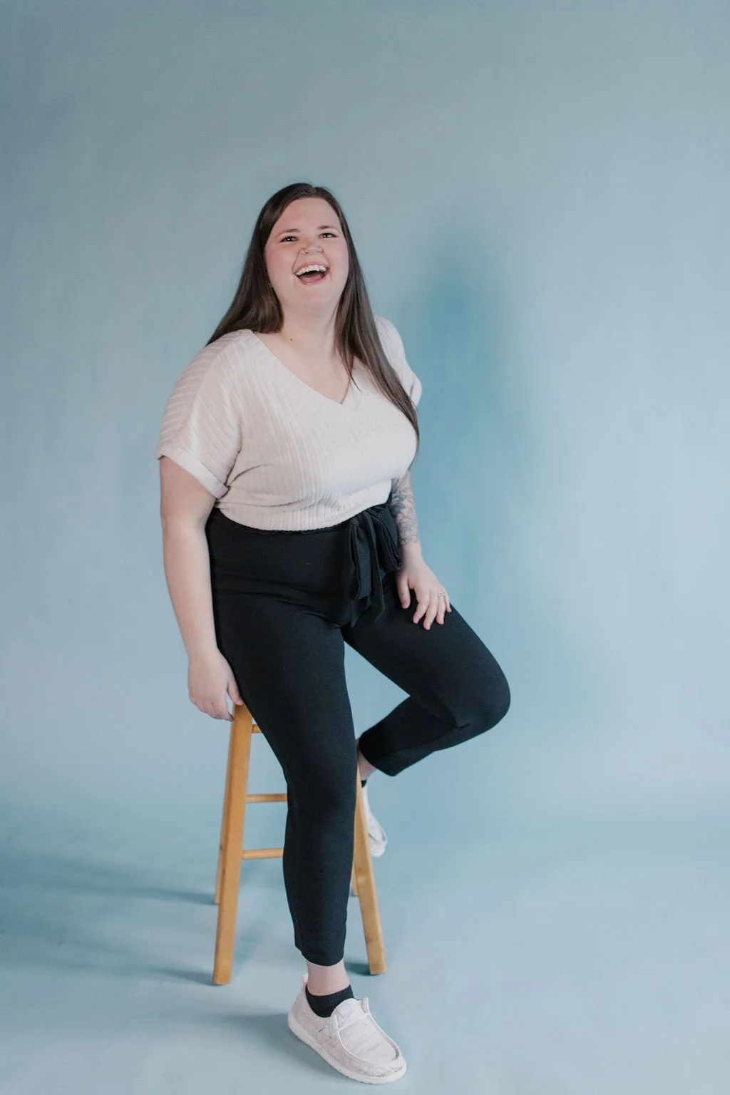 A smiling woman with long brown hair sitting on a wooden stool against a plain light blue background, wearing a beige top, black pants, and white sneakers.