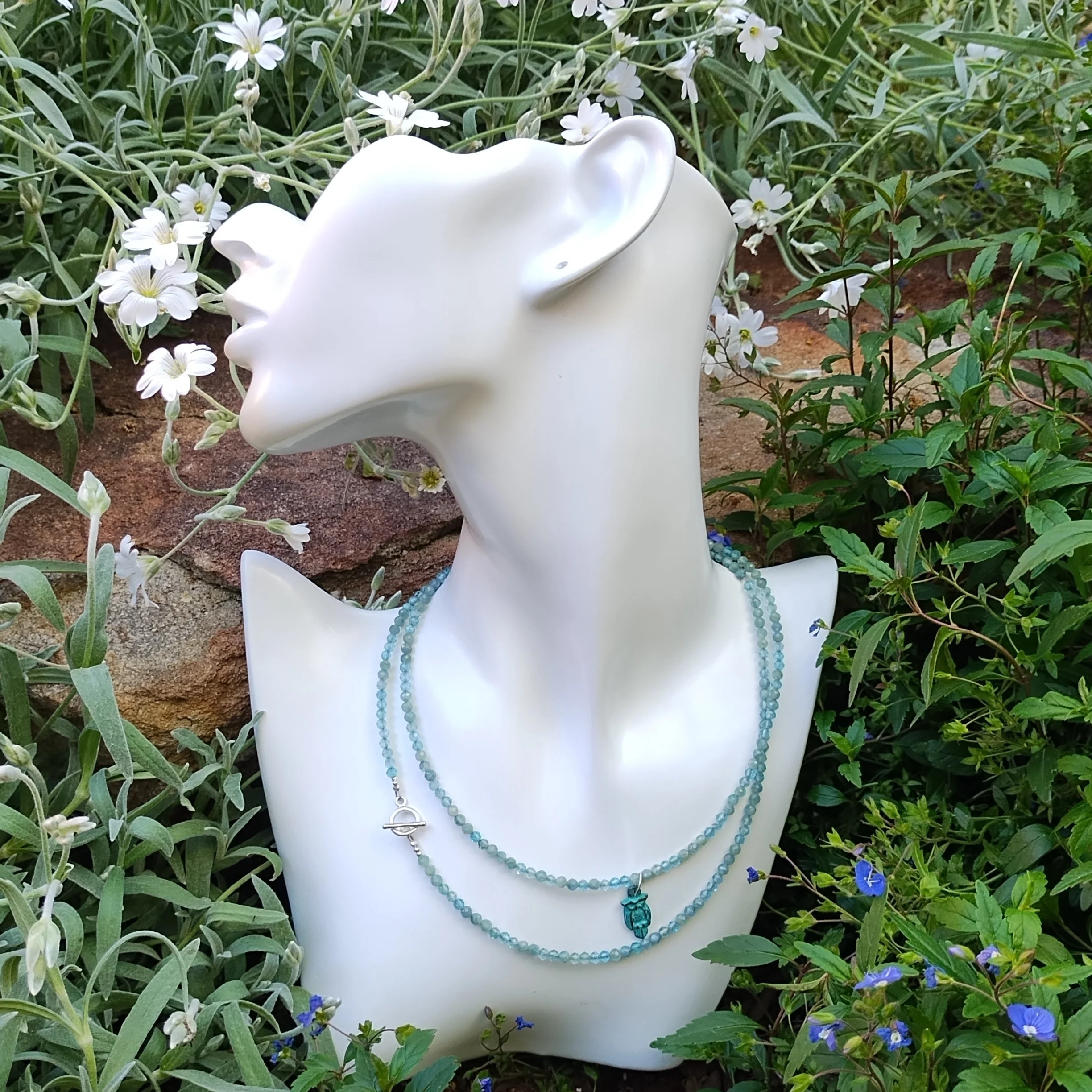White mannequin head adorned with light blue beaded necklaces, surrounded by green foliage and small white and blue flowers.