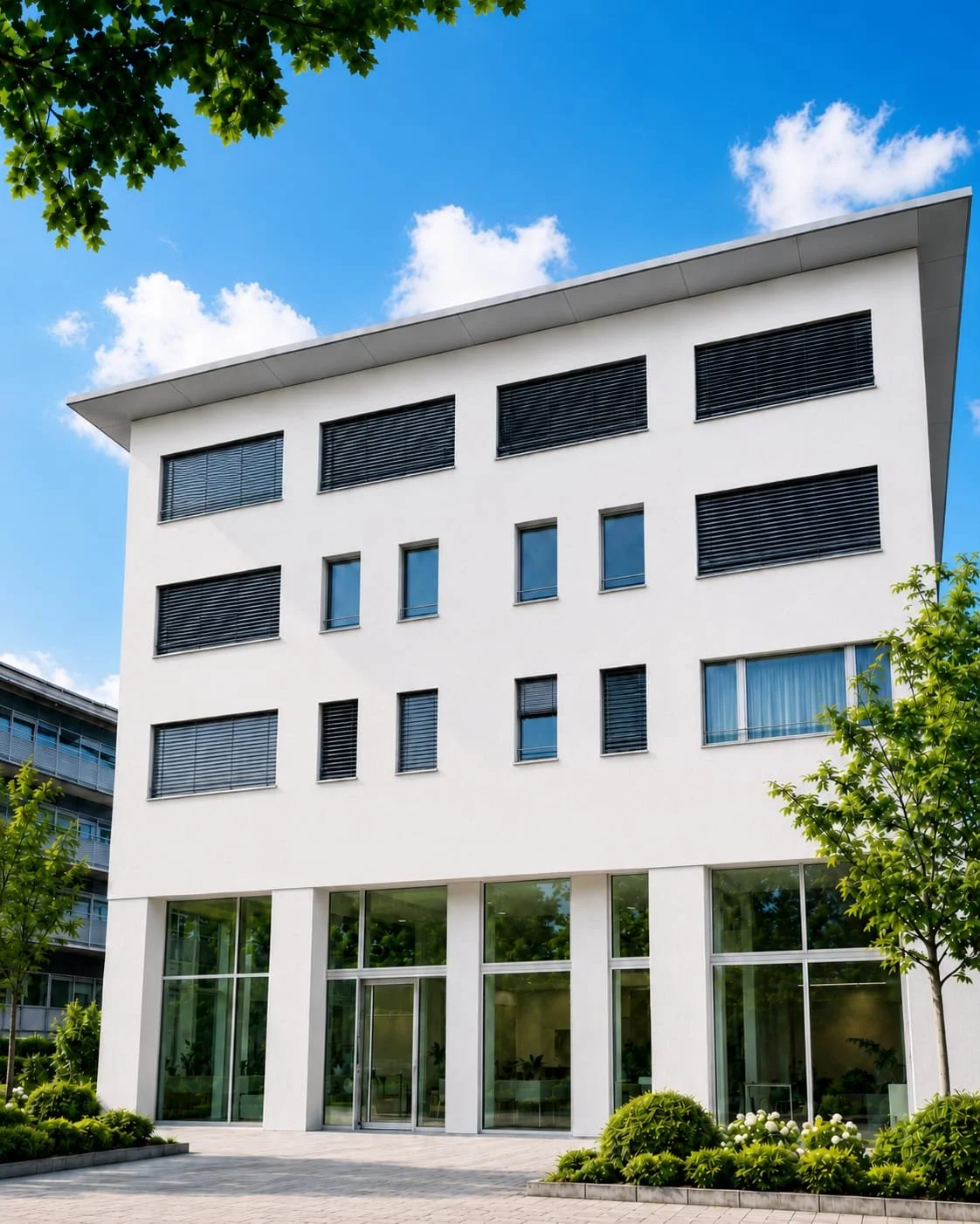 Modernes weißes Mehrfamilienhaus mit großen Fenstern und dunklen Jalousien unter einem blauen Himmel mit Wolken, umgeben von grünen Bäumen.