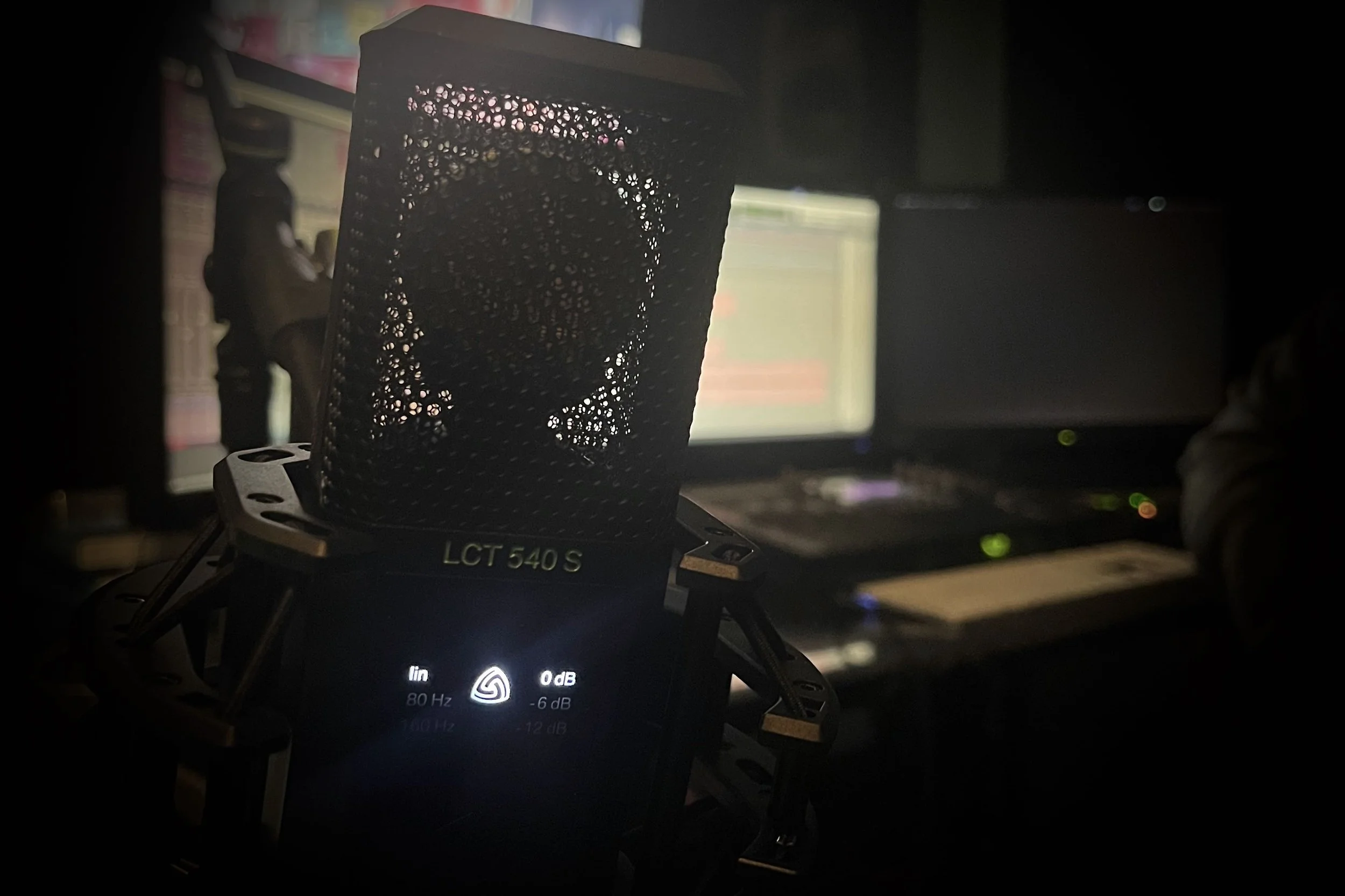 A close-up view of a professional microphone in front of a computer monitor in a dimly lit room.