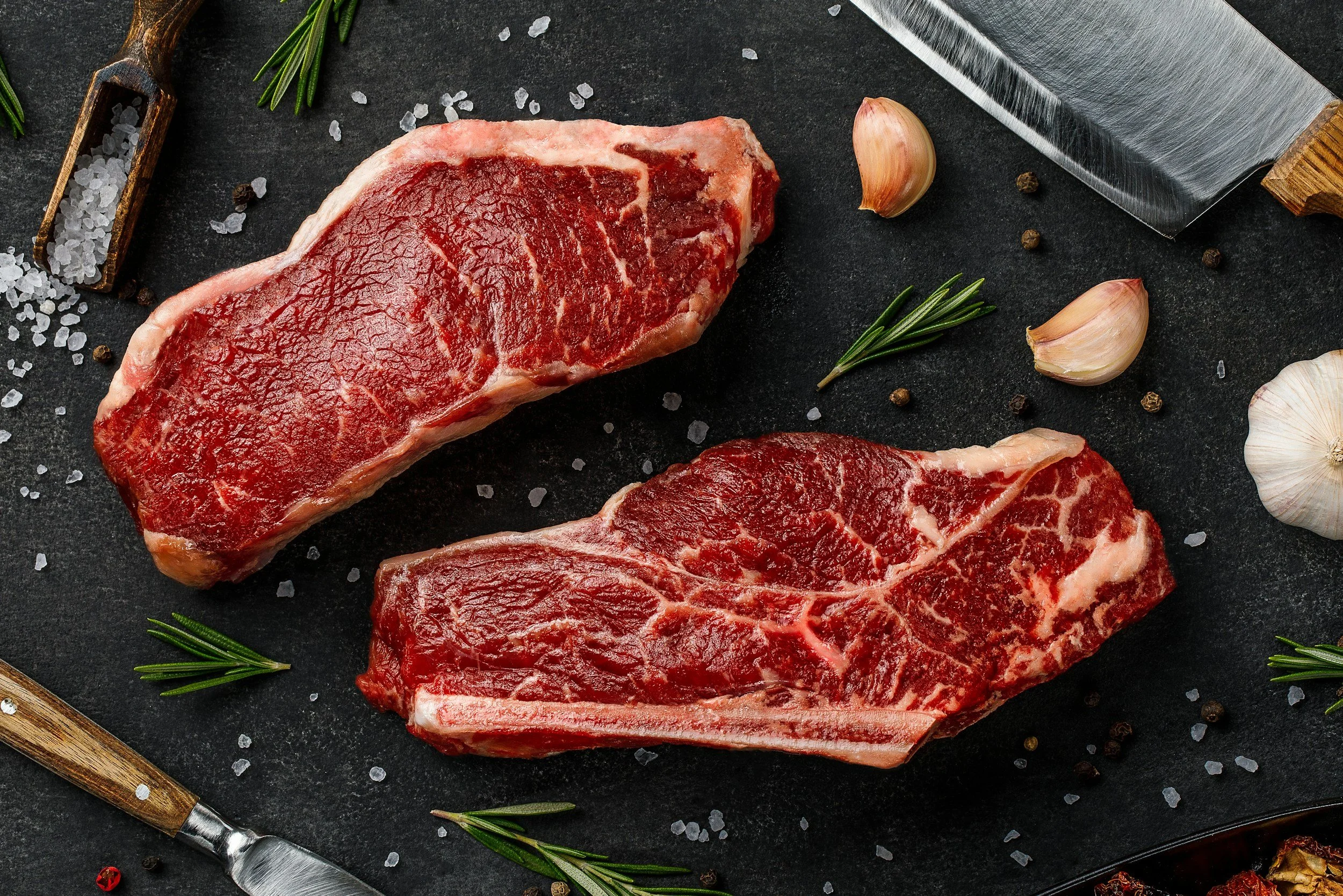 Two raw steaks on a black cutting board with scattered sea salt, garlic cloves, and sprigs of rosemary. A large chef's knife is also visible.