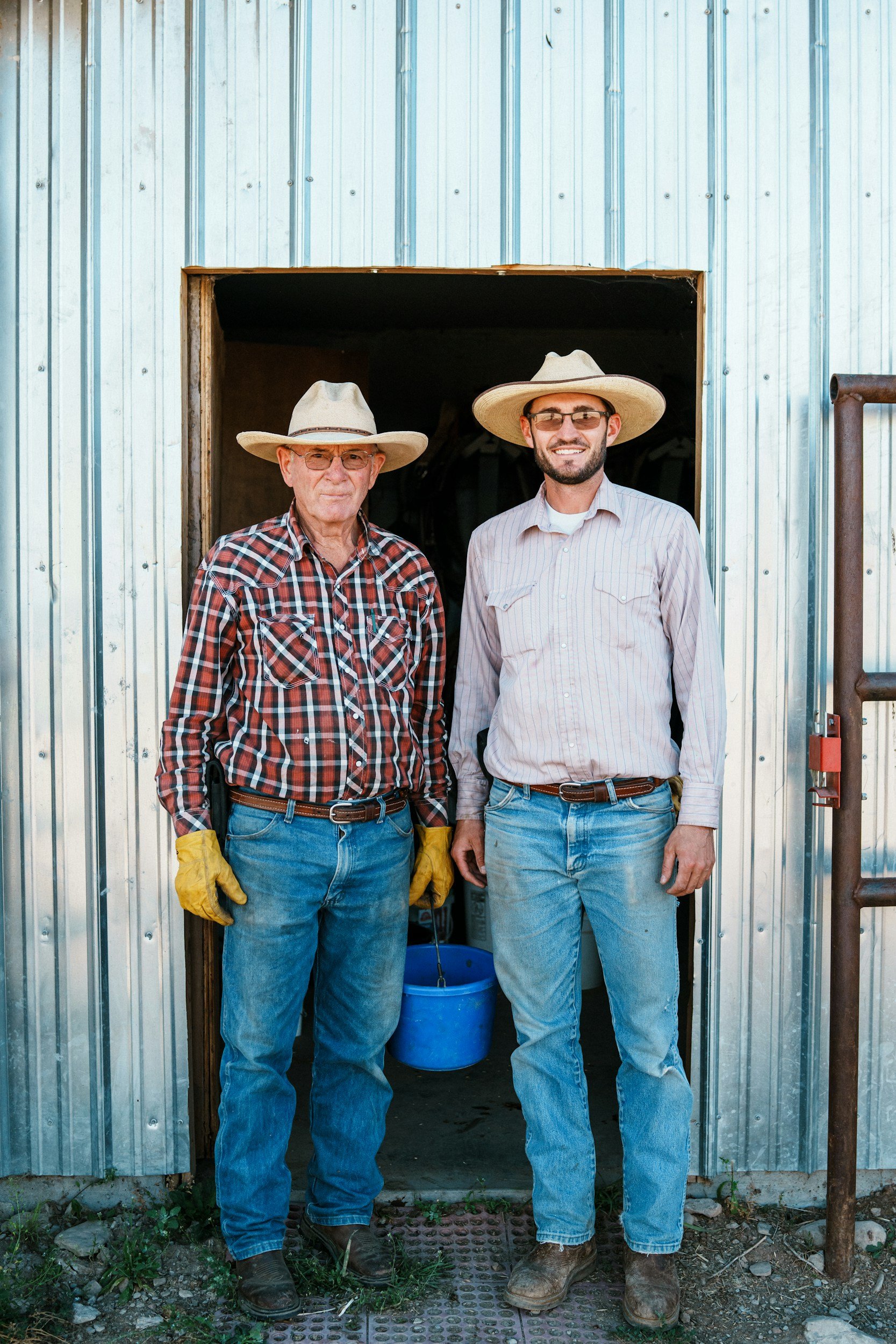 Two men standing in the doorway of a metal building, both wearing cowboy hats, sunglasses, and work clothes. One is elderly and the other is younger, smiling, with a blue bucket behind them.