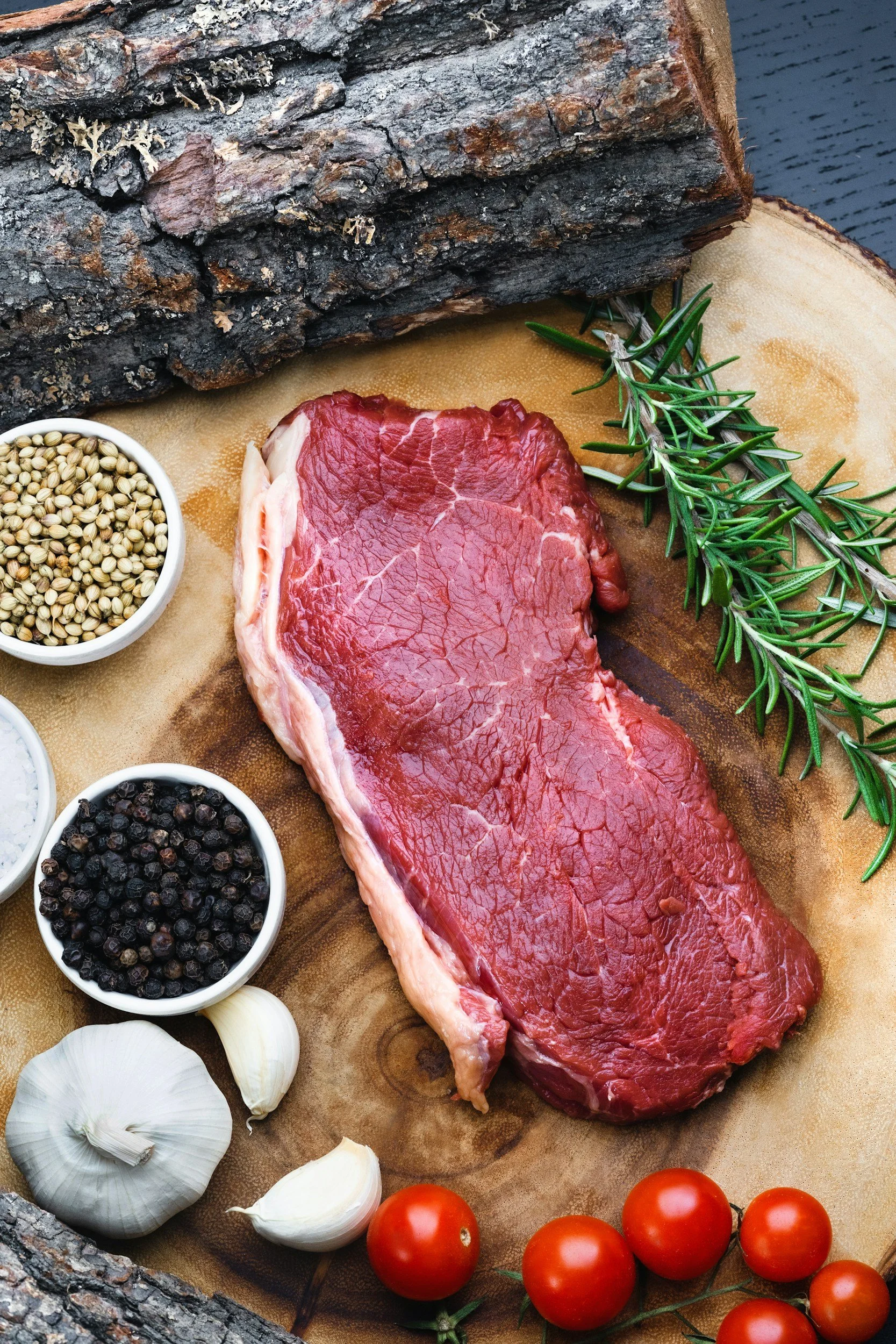 A piece of raw red meat on a wooden cutting board, surrounded by garlic, cherry tomatoes, fresh rosemary, and bowls of peppercorns and coriander seeds, with pieces of wood and a dark background.