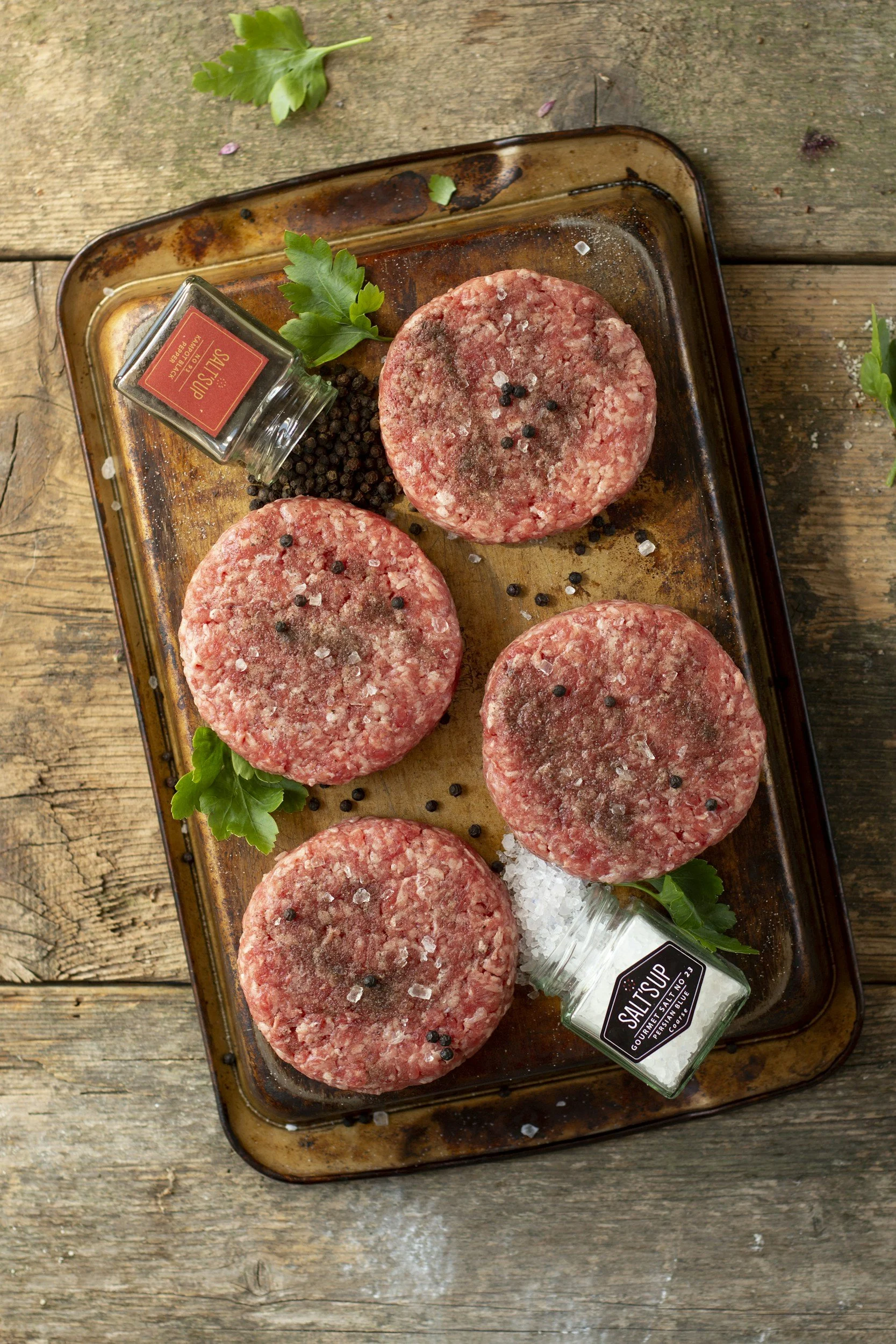 Raw beef burger patties seasoned with salt, black pepper, and sea salt, garnished with fresh parsley on a rusted baking sheet, on a wooden surface.