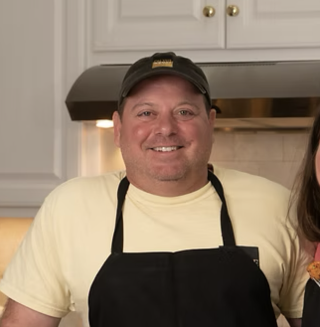Smiling man in a yellow t-shirt wearing a black apron and cap in a kitchen.