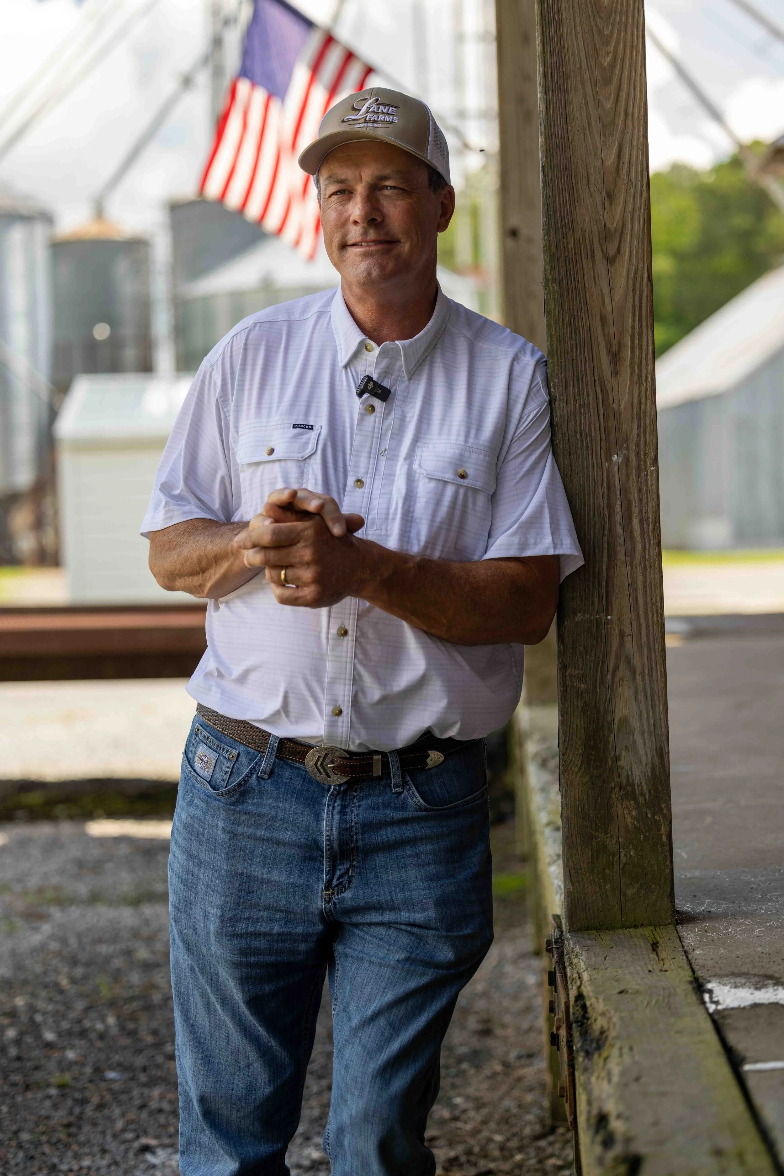A man wearing a gray cap, white button-up shirt, and jeans stands outdoors leaning against a wooden post, with an American flag visible in the background.